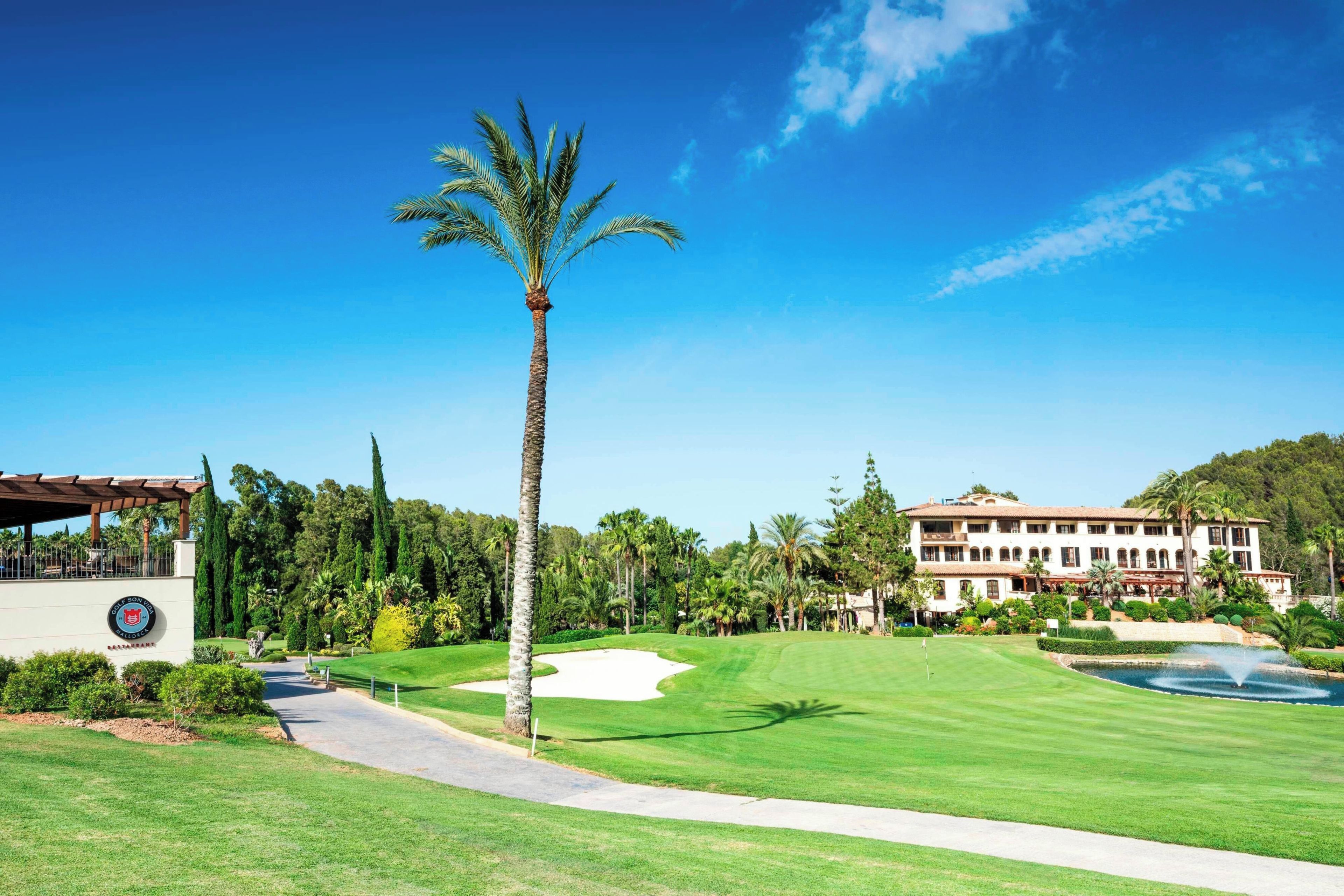 A golf course with a clubhouse, palm trees, and a large building in the background.
