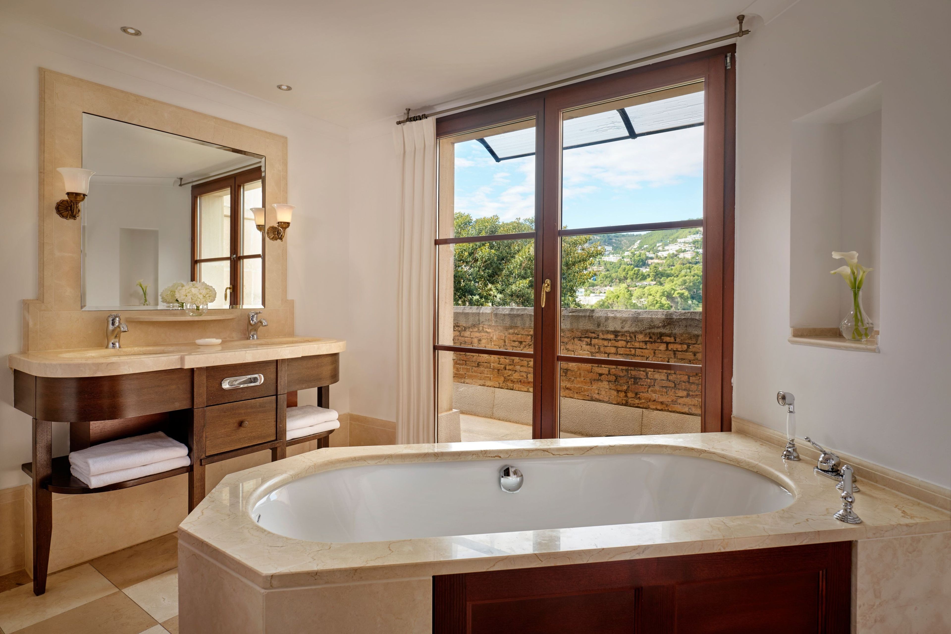 Une salle de bain avec une grande baignoire, un meuble-lavabo avec un miroir et deux lampes, une fenêtre donnant sur un paysage verdoyant et une étagère avec un vase.