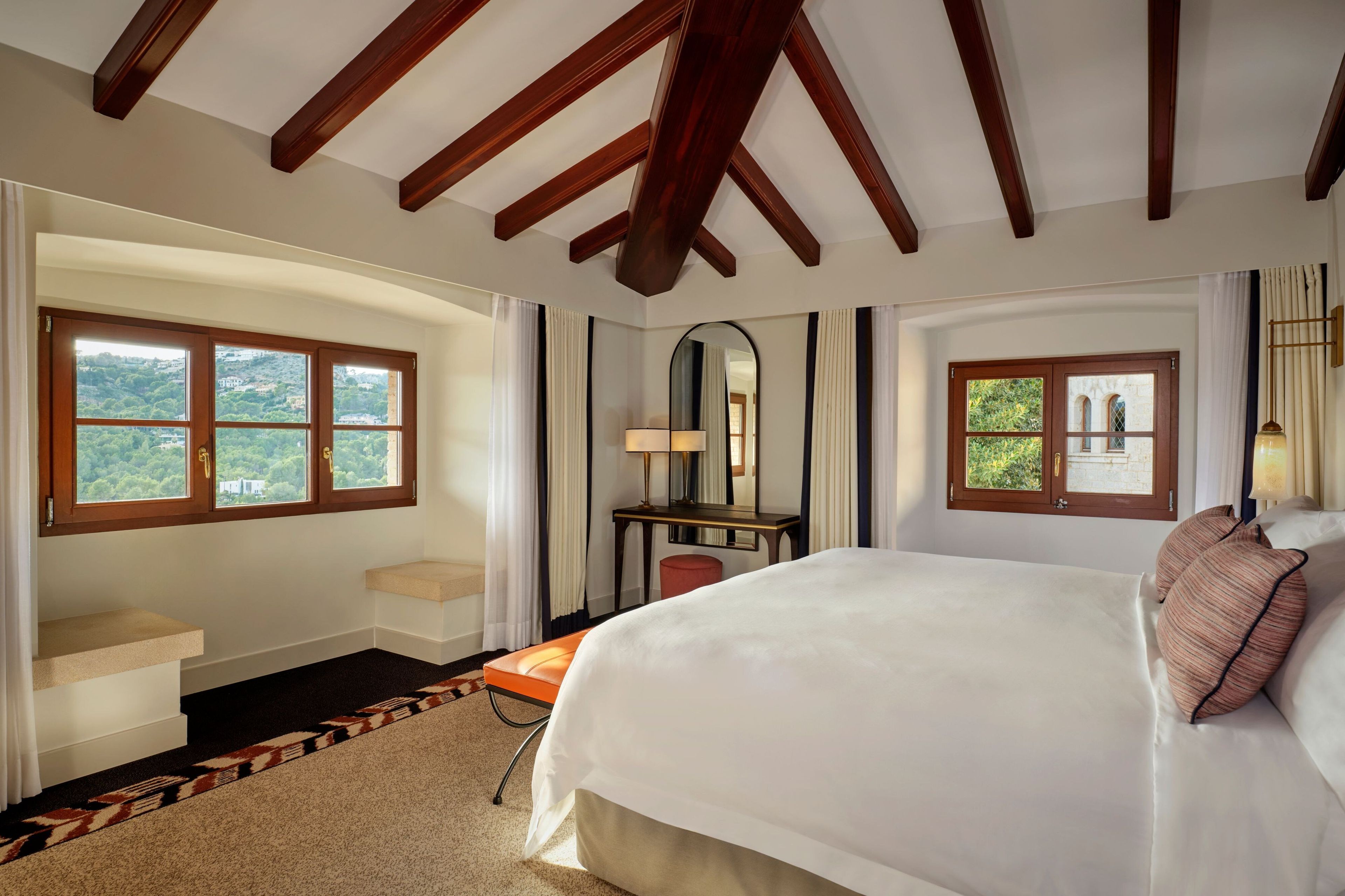 Habitación de hotel con una cama grande, un escritorio y una silla. Hay dos ventanas con cortinas, un espejo y vistas a zonas verdes.