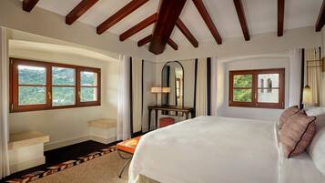 Habitación de hotel con una cama grande, un escritorio y una silla. Hay dos ventanas con cortinas, un espejo y vistas a zonas verdes.
