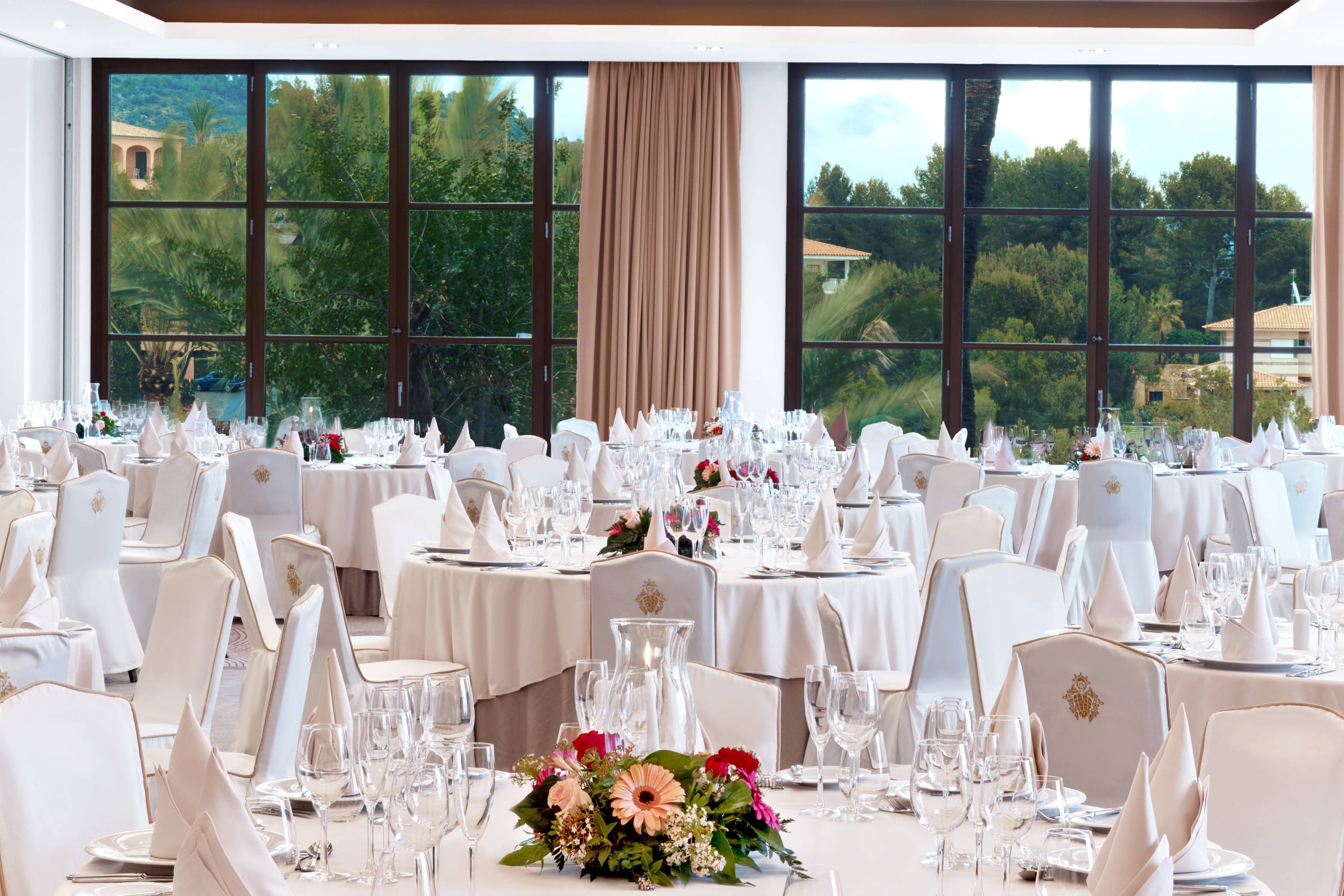 Un salón de banquetes con mesas redondas dispuestas para un evento formal, grandes ventanales con vistas a zonas verdes y edificios, y manteles y sillas blancos.