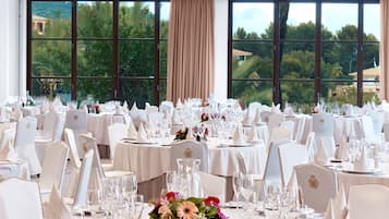 Un salón de banquetes con mesas redondas dispuestas para un evento formal, grandes ventanales con vistas a zonas verdes y edificios, y manteles y sillas blancos.