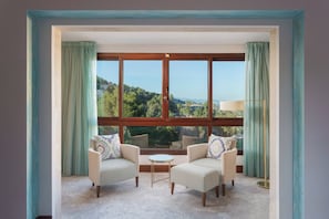 Ein Zimmer mit zwei Sesseln und einem kleinen Tisch, einem großen Fenster mit Vorhängen und Blick auf eine grüne Landschaft.