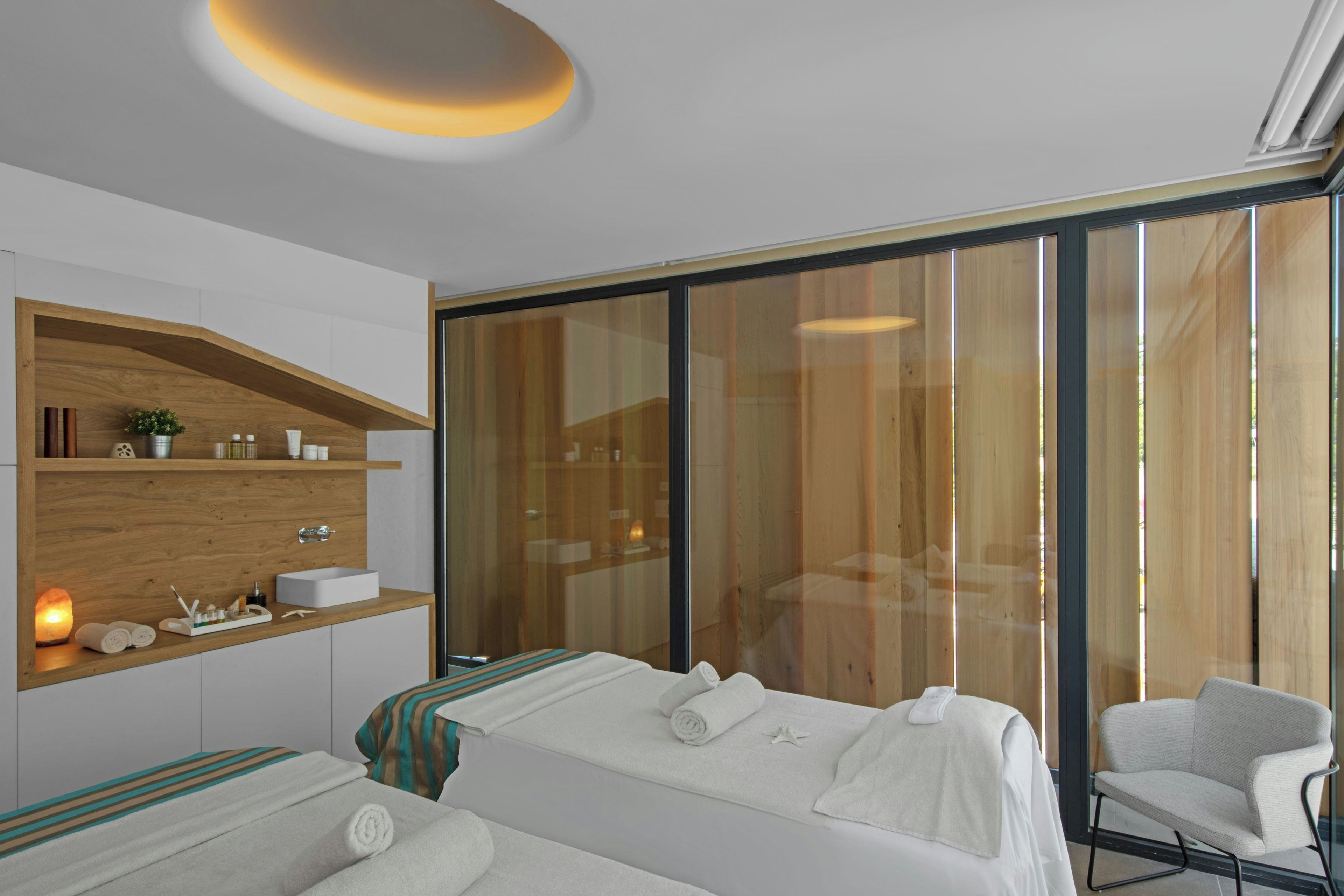A modern hotel room with two beds, a wooden shelf with toiletries, a glass shower enclosure, and a seating area.