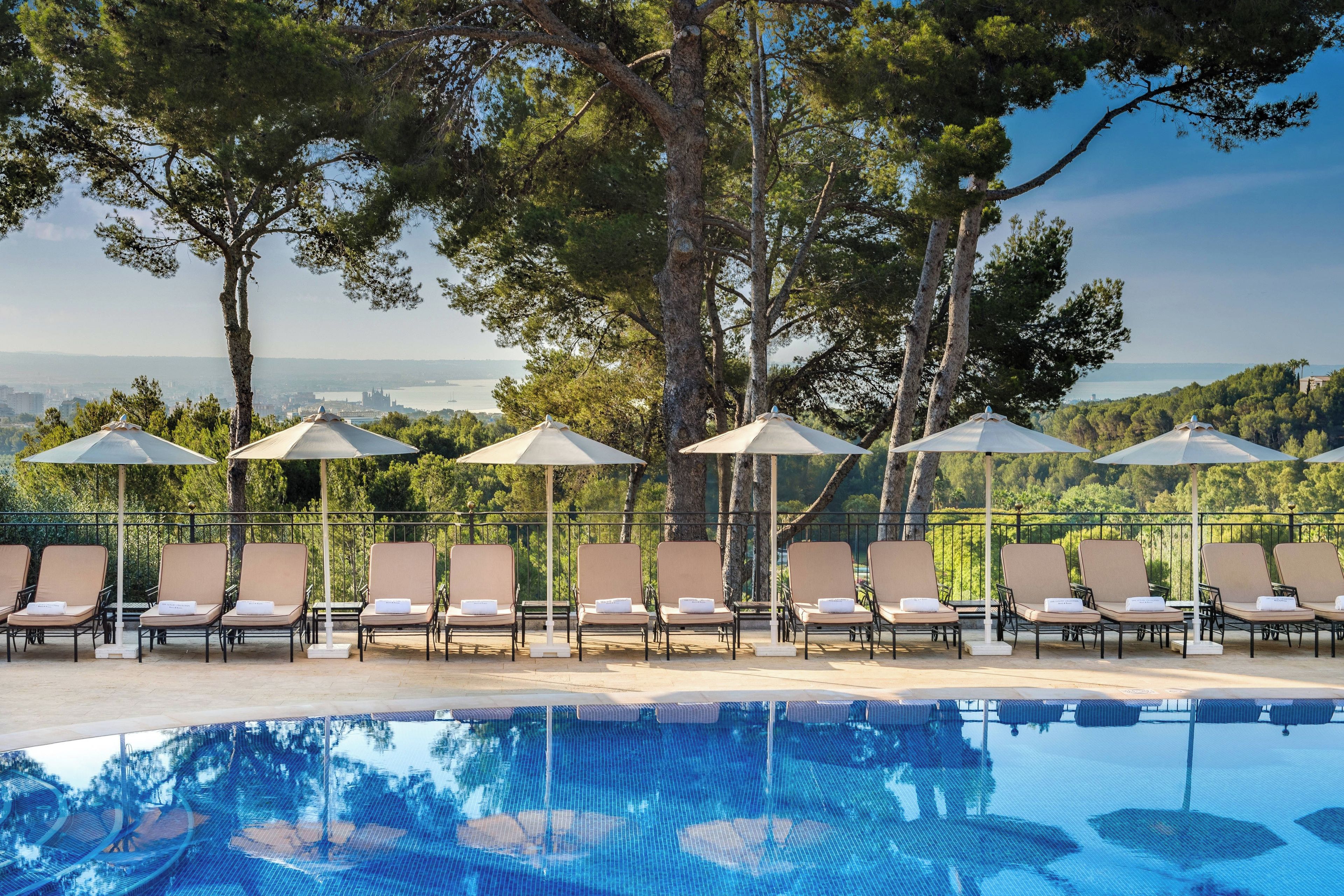 A pool area with lounge chairs and umbrellas, surrounded by trees and overlooking a body of water.