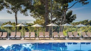 A pool area with lounge chairs and umbrellas, surrounded by trees and overlooking a body of water.