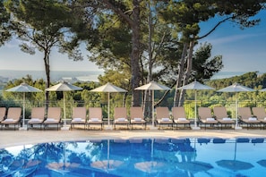 A pool area with lounge chairs and umbrellas, surrounded by trees and overlooking a body of water.