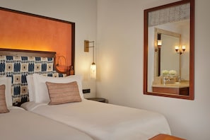 Habitación de hotel con cama, mesita de noche, lámpara de pared, espejo con marco decorativo y baño con lavamanos y espejo.