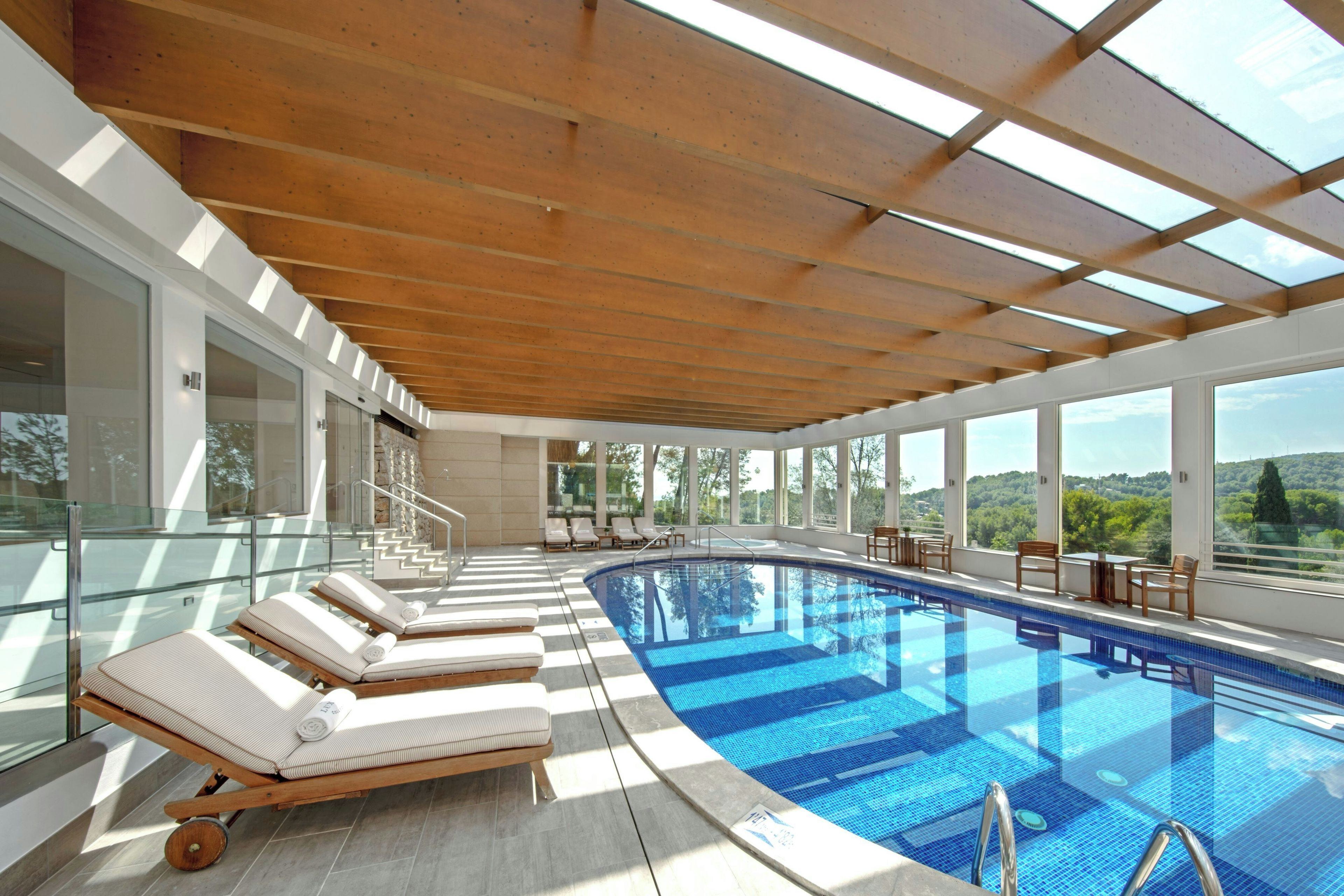 A modern indoor swimming pool with a wooden ceiling and glass walls, offering views of the surrounding landscape.