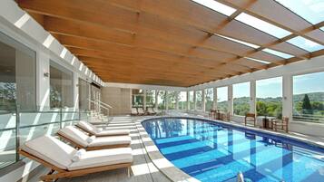 A modern indoor swimming pool with a wooden ceiling and glass walls, offering views of the surrounding landscape.