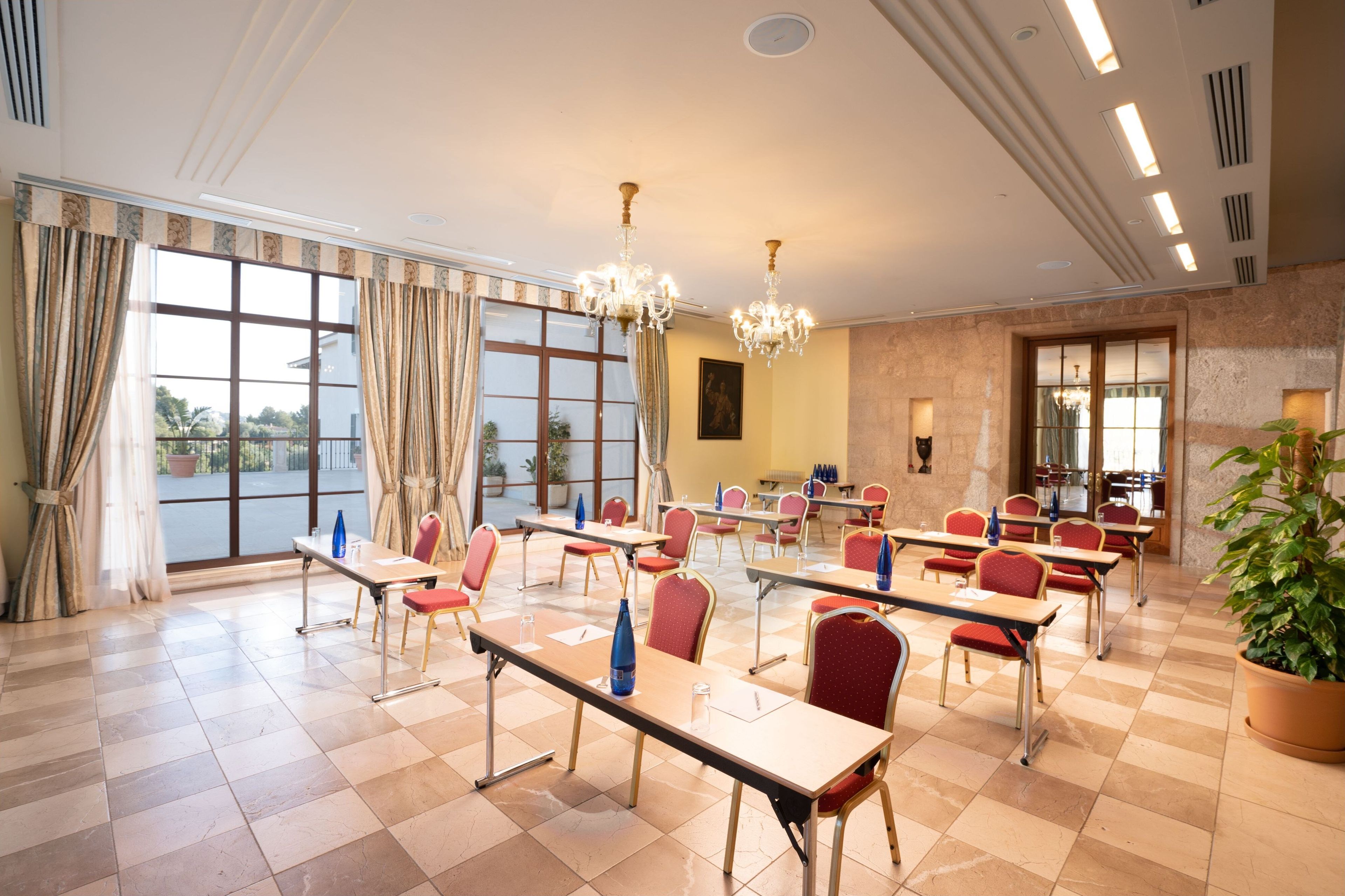 Een congreszaal met meerdere tafels en stoelen, grote ramen met gordijnen, kroonluchters en een potplant.