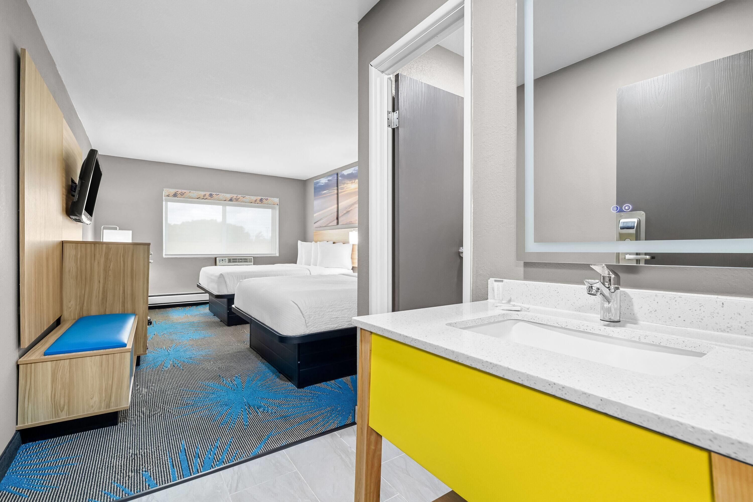 Une chambre d’hôtel comprenant un lit, un petit coin salon, une salle de bain avec un lavabo et un miroir, et une grande fenêtre.