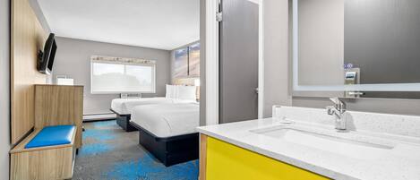 Une chambre d’hôtel comprenant un lit, un petit coin salon, une salle de bain avec un lavabo et un miroir, et une grande fenêtre.