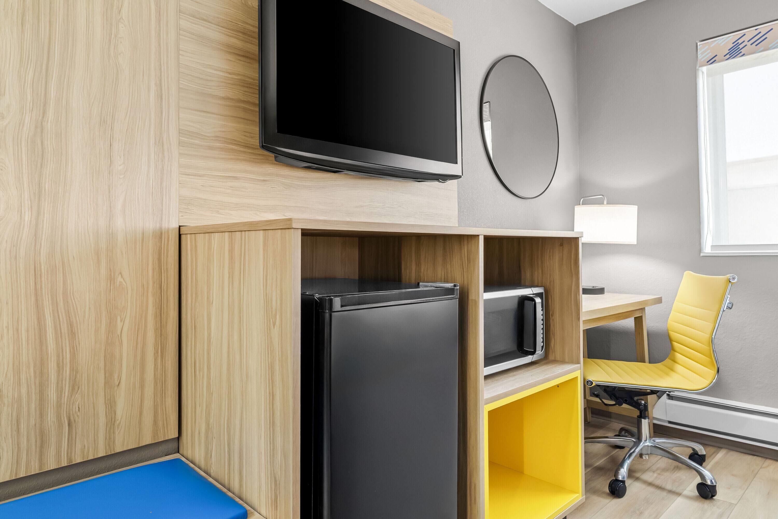 A modern room with a flat-screen TV, a refrigerator, a microwave, a yellow office chair, and a desk.