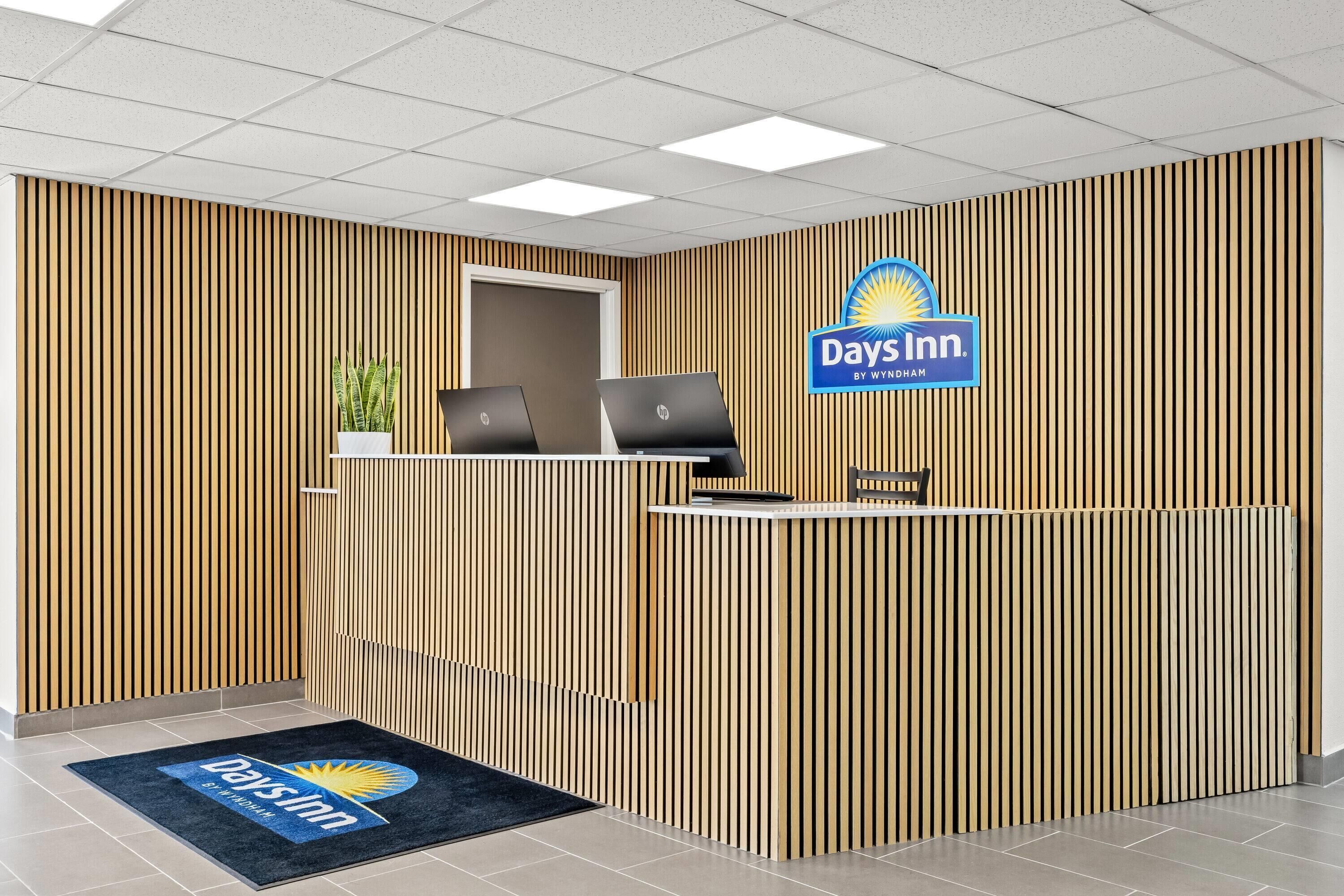 Un comptoir d’accueil orné du logo de Days Inn, deux ordinateurs portables, une plante en pot et un paillasson bleu.