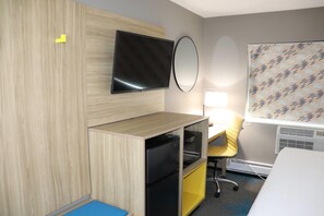 Une chambre d’hôtel équipée d’un téléviseur à écran plat, d’un miroir, d’un bureau avec une chaise, d’un petit réfrigérateur et d’un store à motifs.