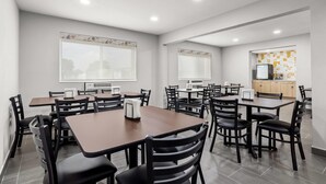 A modern dining area with multiple tables and chairs, a kitchen area with appliances, and large windows with patterned blinds. - Days Inn by Wyndham La Crosse (La Crosse)