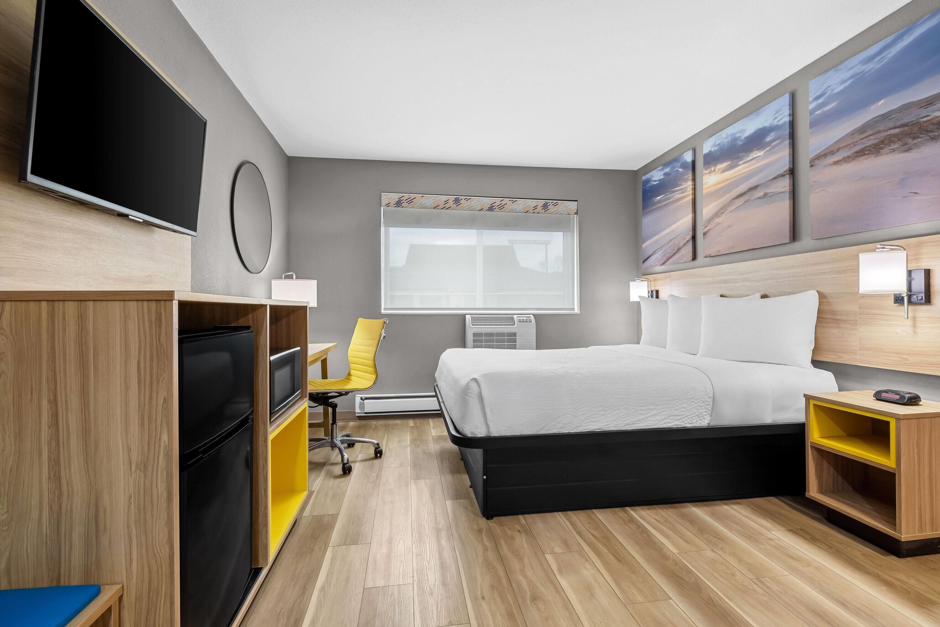 A hotel room with a bed, a TV, a desk, a chair, and artwork on the wall.