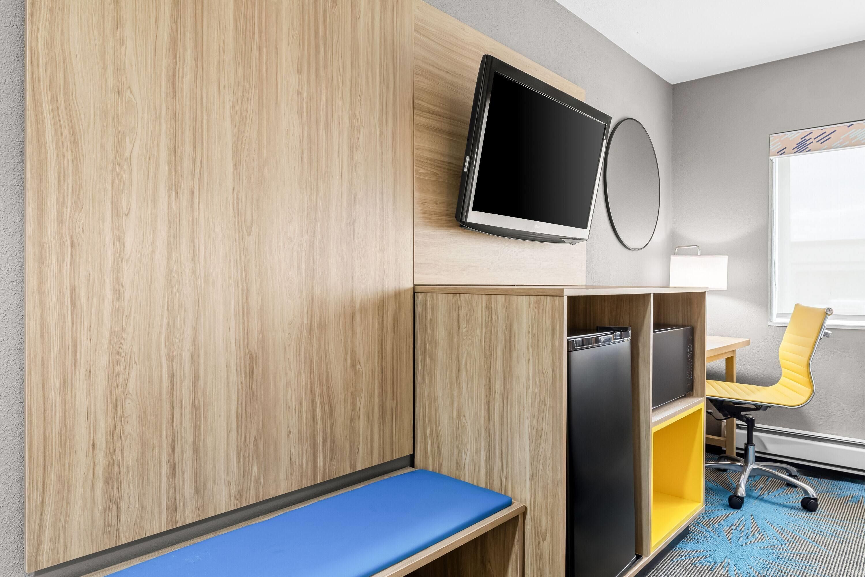 Une chambre d’hôtel avec une armoire en bois, un banc bleu, une chaise jaune, un téléviseur et une petite table.