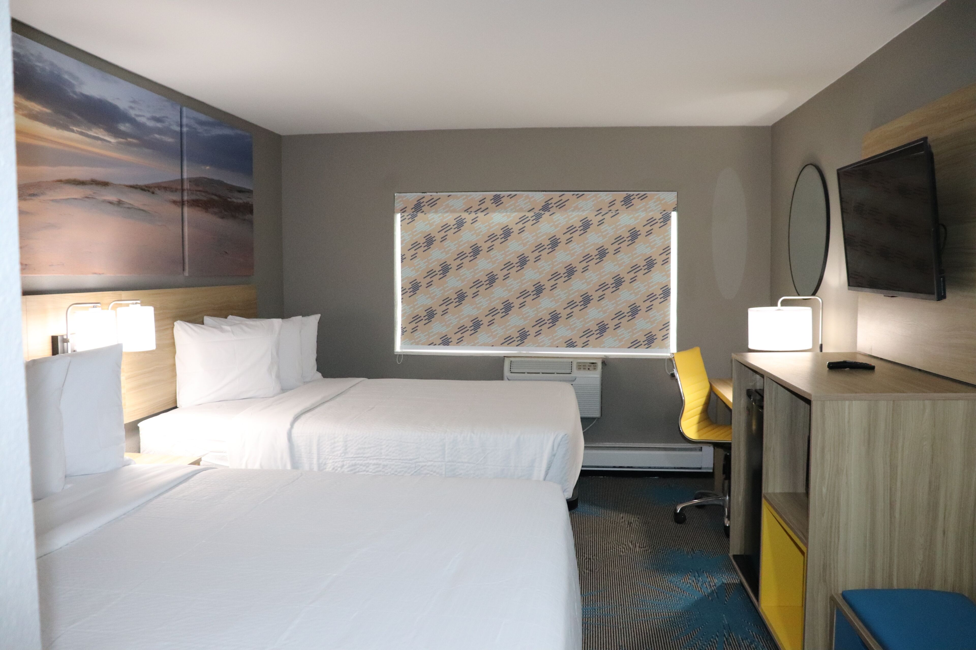 A hotel room with two beds, a desk, a chair, a TV, and a window with a patterned curtain.