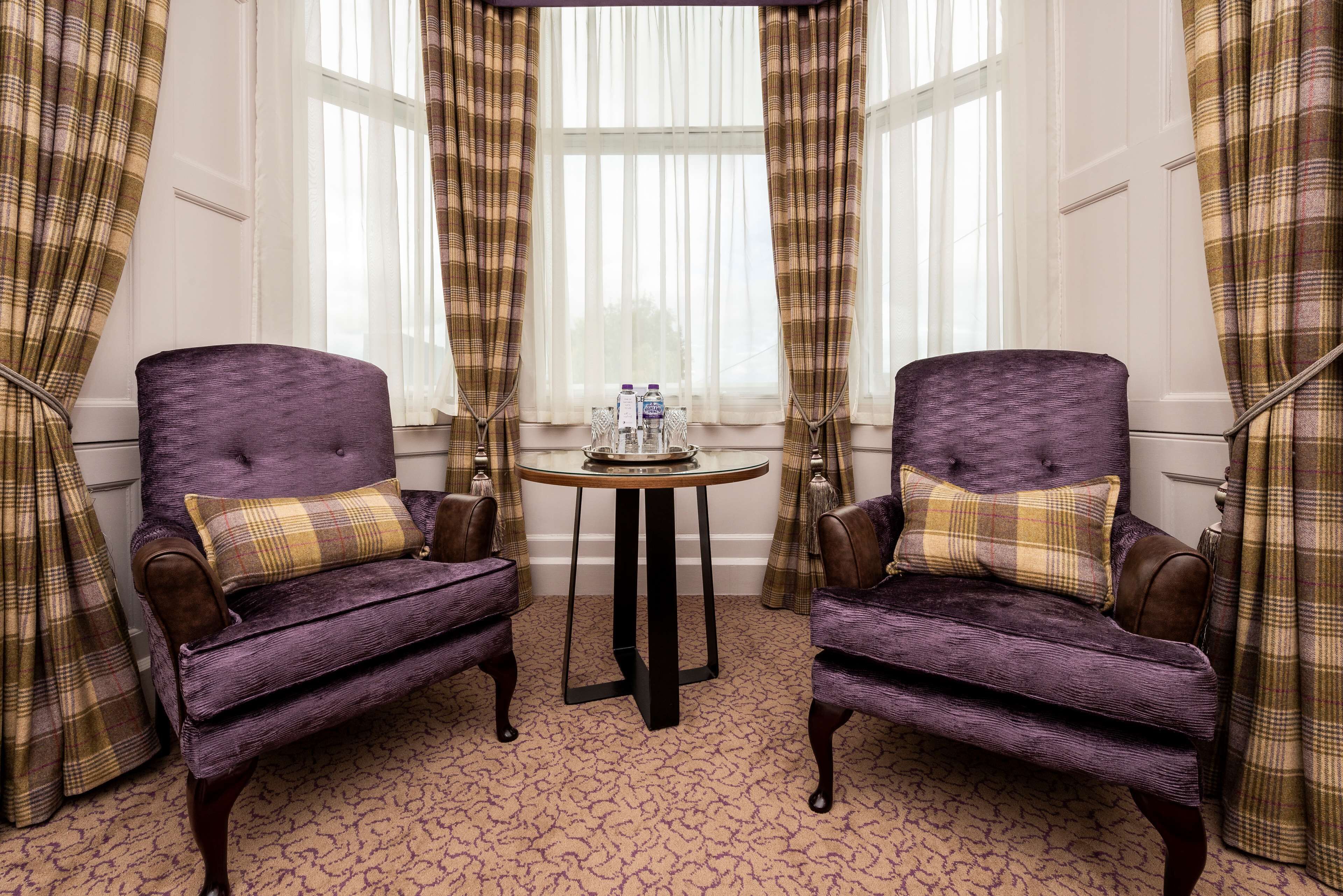 Deux fauteuils violets avec des coussins à carreaux, une table ronde sur laquelle se trouve un plateau rempli d’objets, et des rideaux à carreaux.