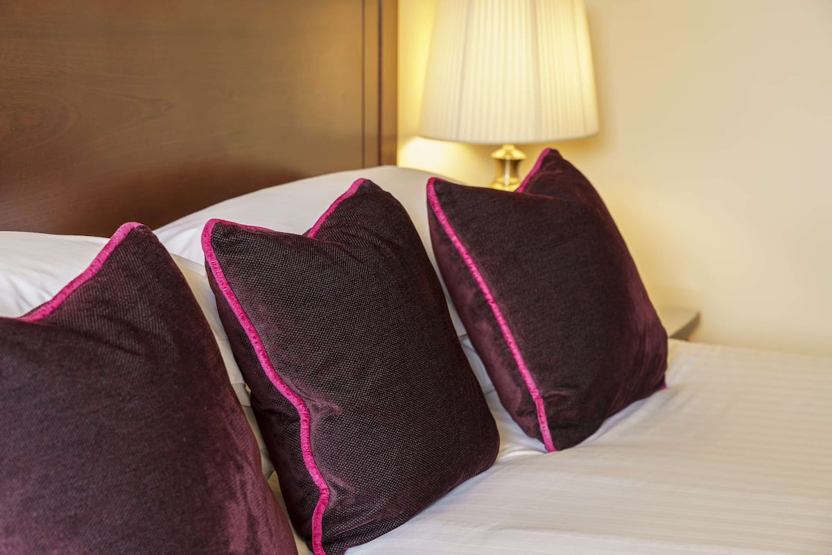 A bed with three dark pillows and a white pillow, a lamp, and a wooden headboard.