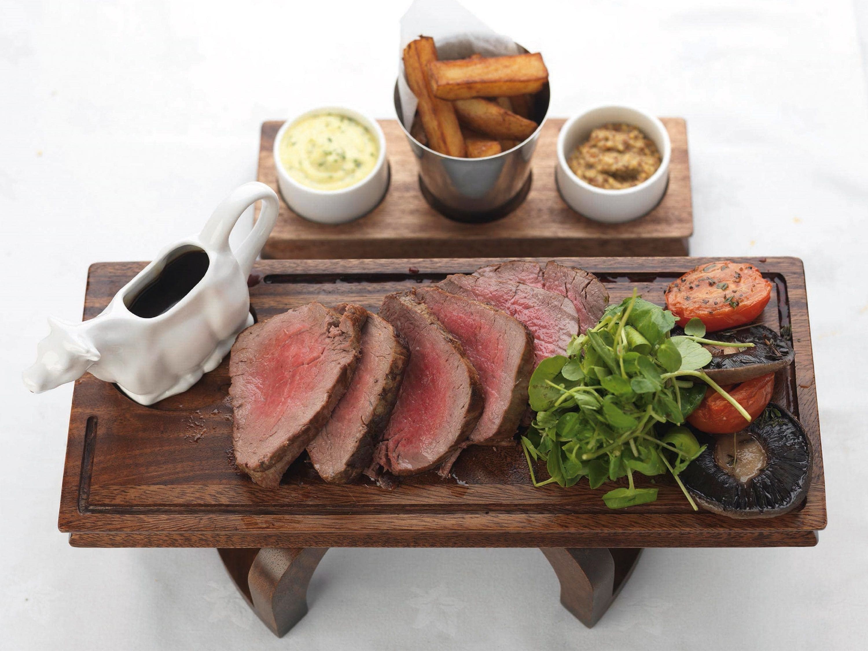 A wooden board with sliced beef, roasted vegetables, and two small bowls of sauces.