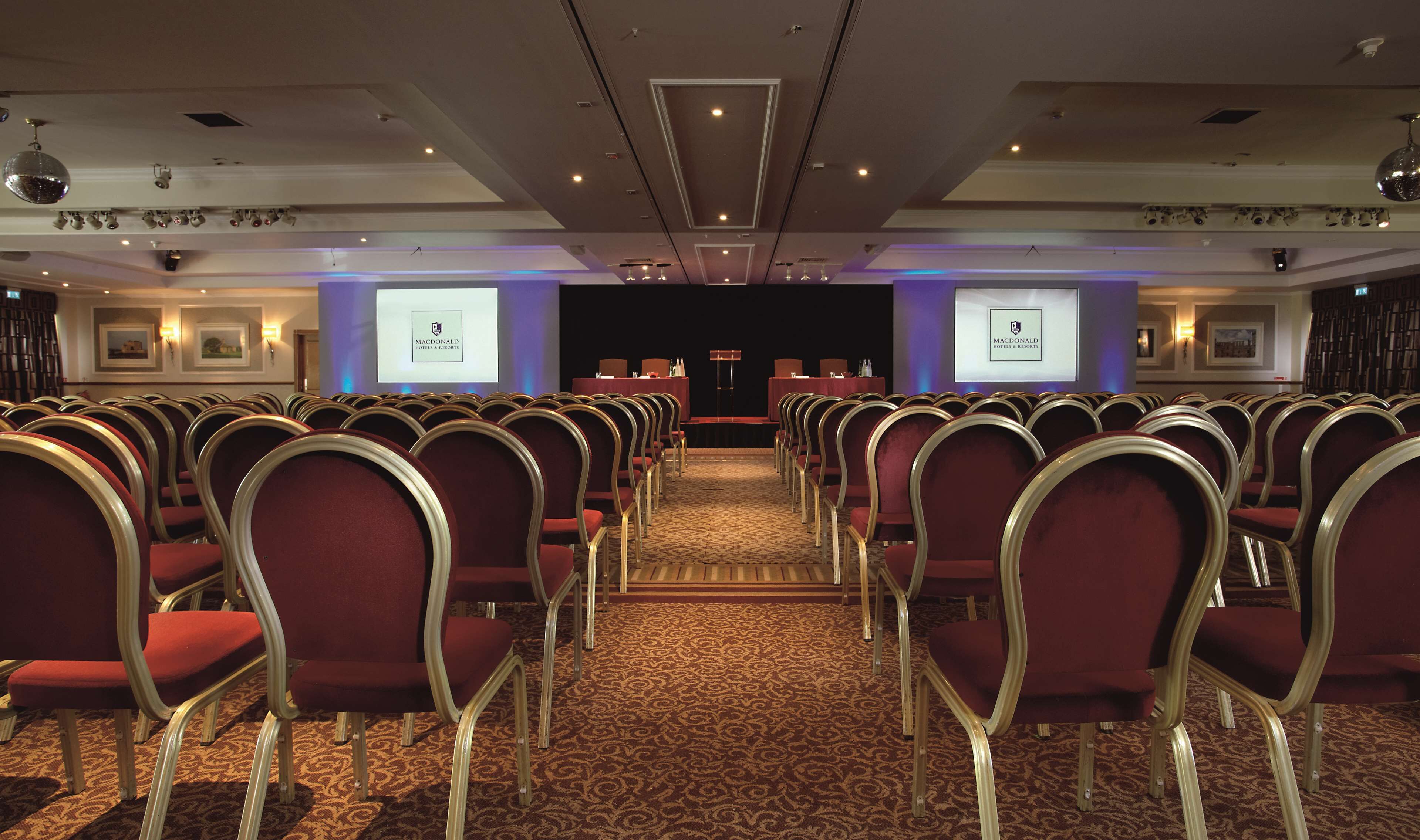 Un salón de conferencias con filas de sillas rojas, un atril y dos pantallas mostrando el logotipo de Macdonald Hotels & Resorts.
