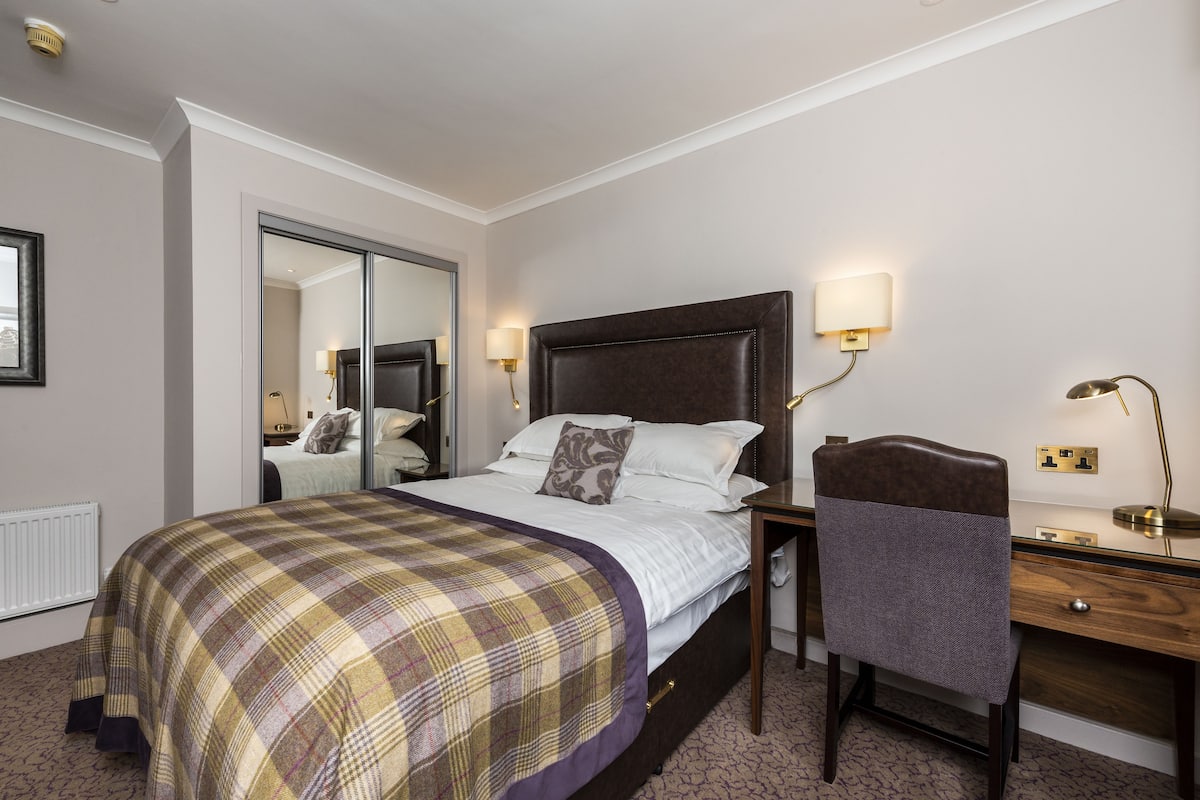 A hotel room with a large bed, a desk with a chair, a mirror, and bedside lamps.