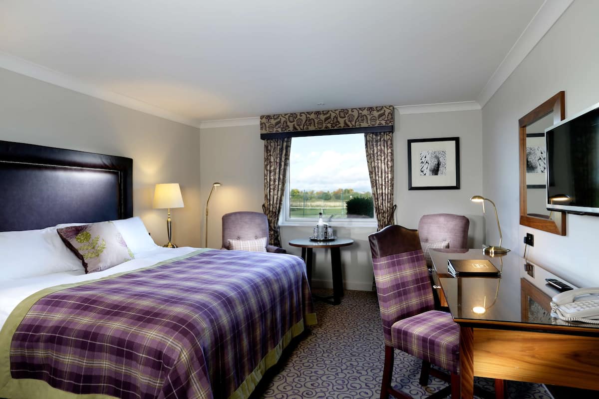 A hotel room with a bed, two chairs, a desk, a lamp, a telephone, and a window with curtains.