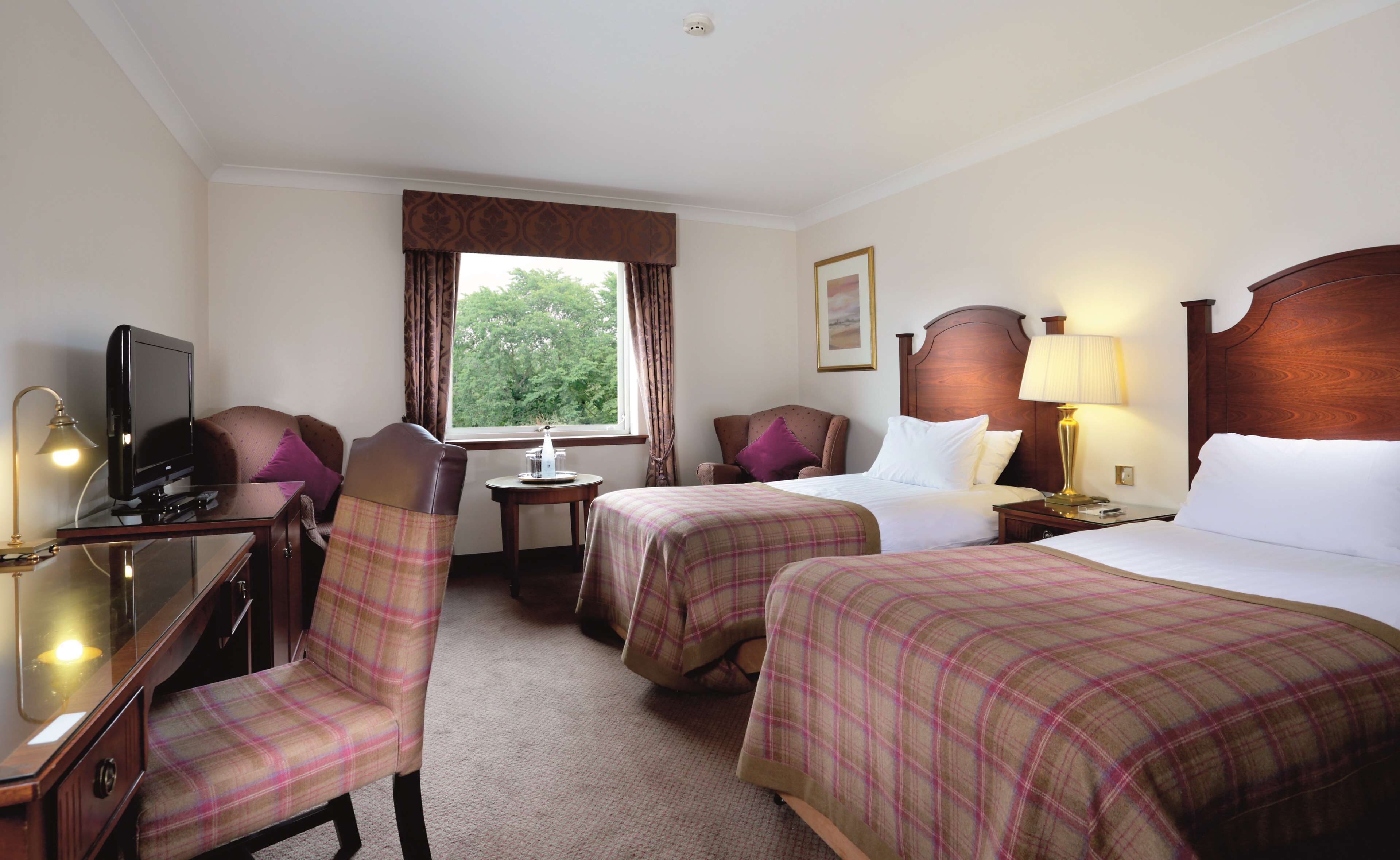 A hotel room with two beds, a desk with a television, a chair, a lamp, and a window with curtains.