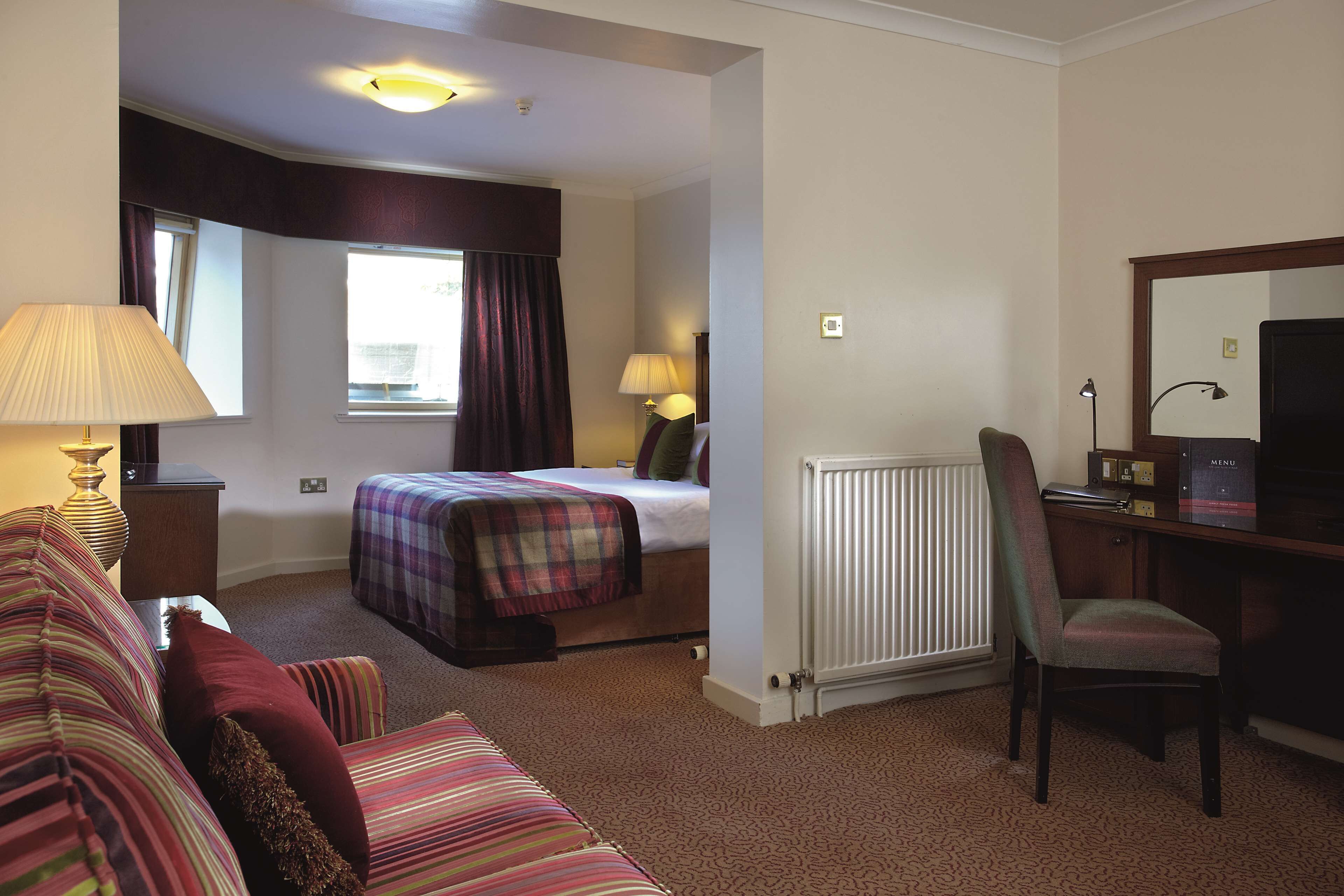 A hotel room with a bed, a desk, a chair, a lamp, and a window with curtains.