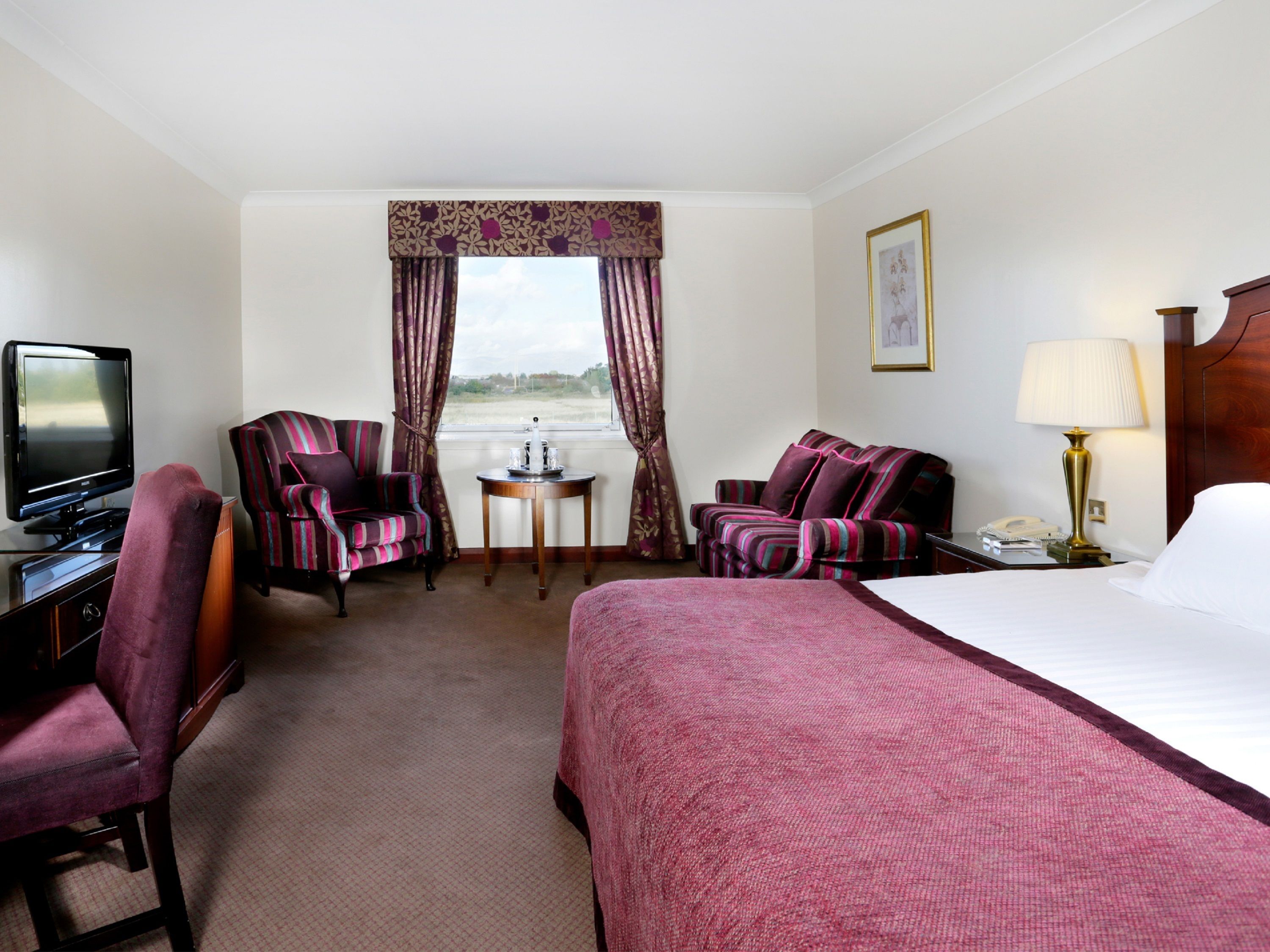 A hotel room with a bed, two armchairs, a desk with a television, a bedside table with a lamp, and a window with curtains.