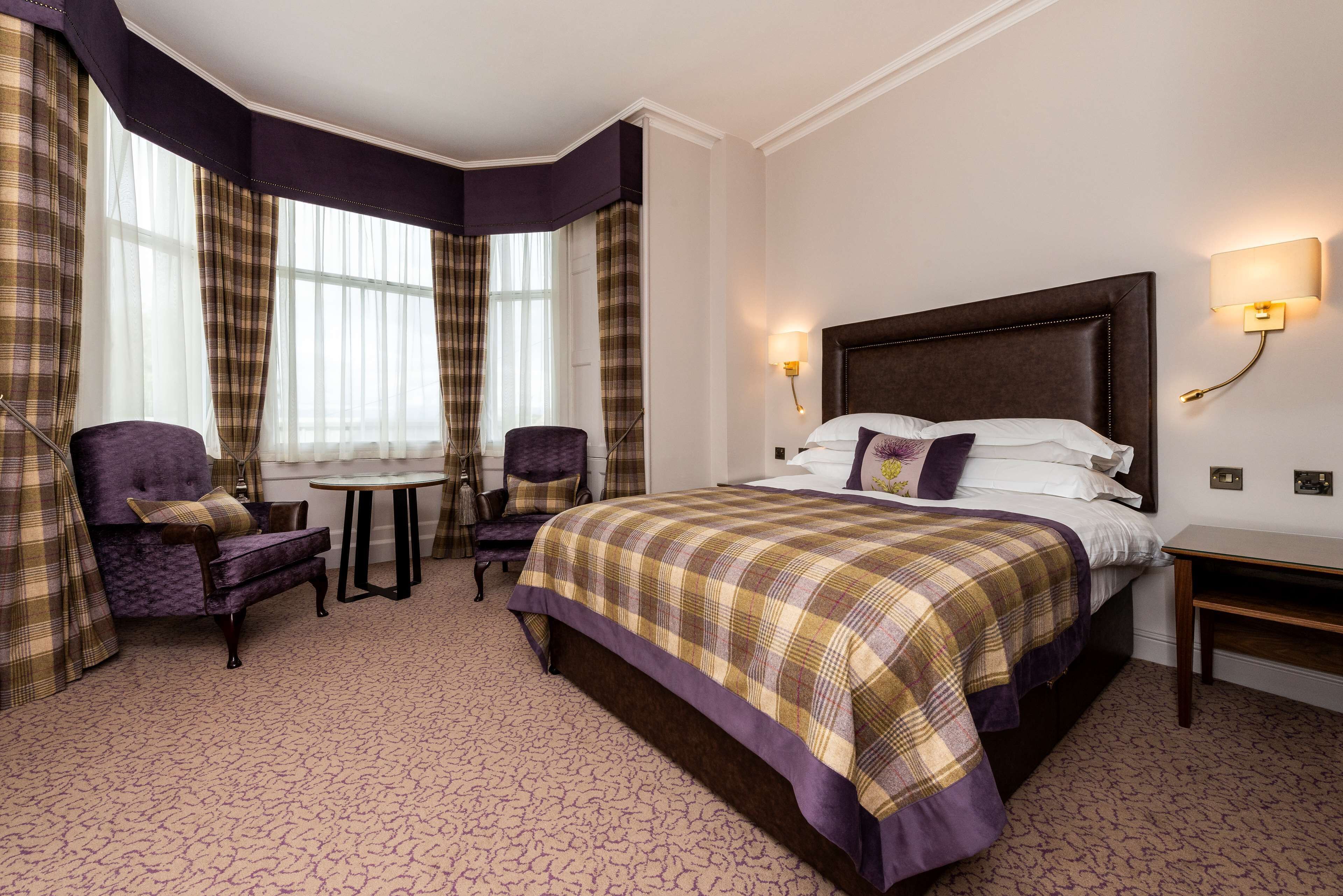 A hotel room with a large bed, two armchairs, a small table, and a window with curtains.