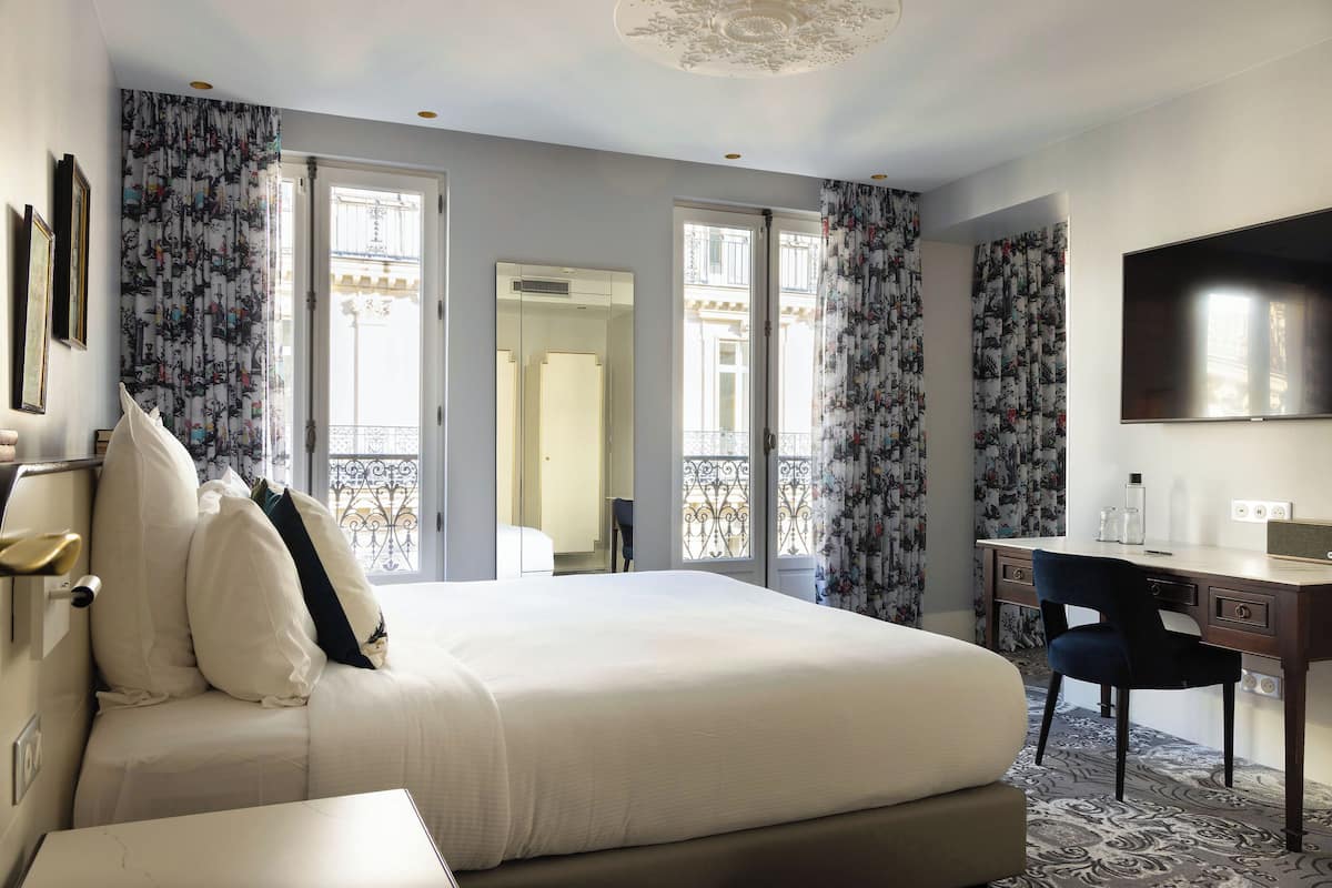 A hotel room with a large bed, a desk, and a chair. There is a TV on the wall and a window with curtains.