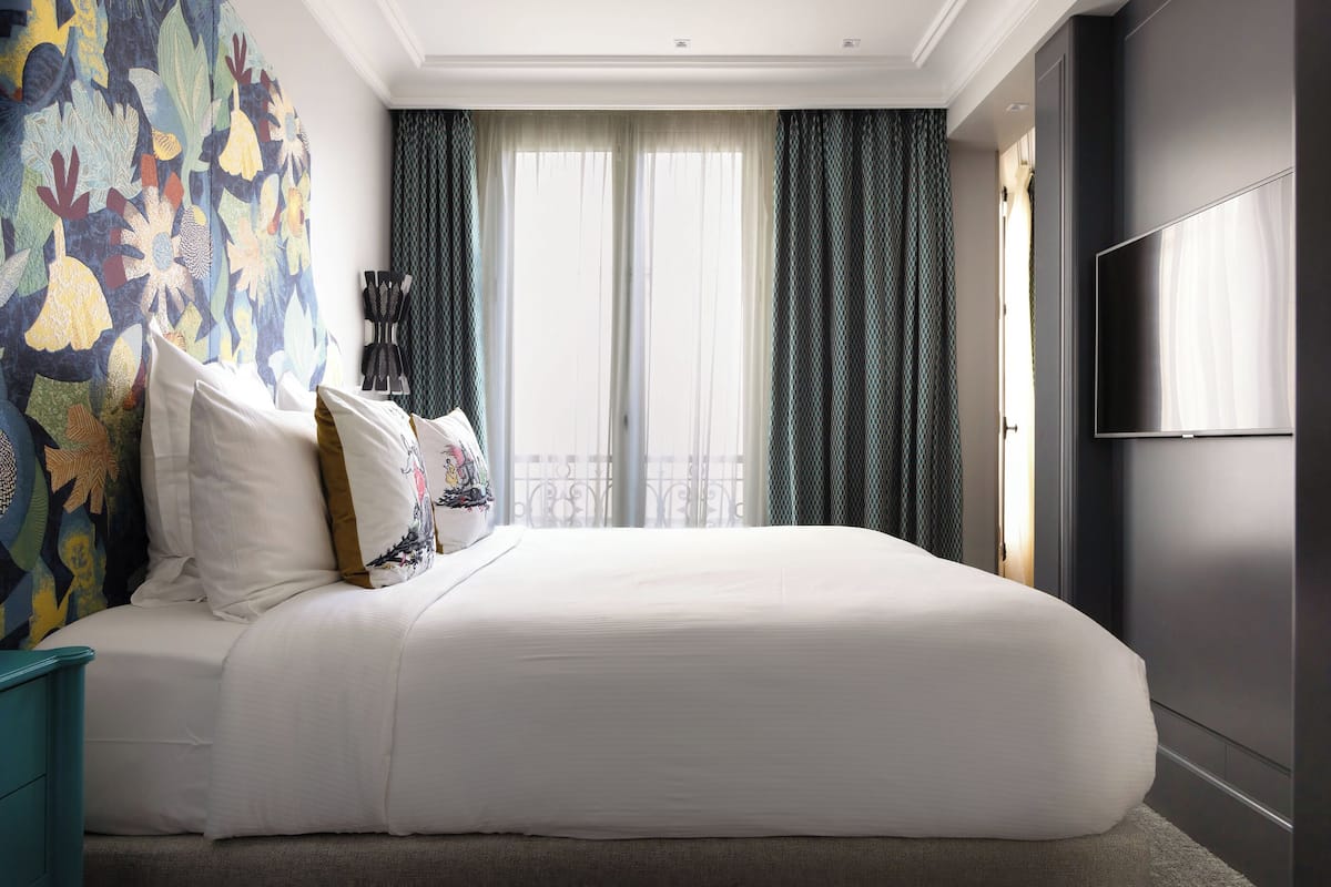 A large bed with white linens, a colorful abstract wall mural, and a large window with sheer curtains.
