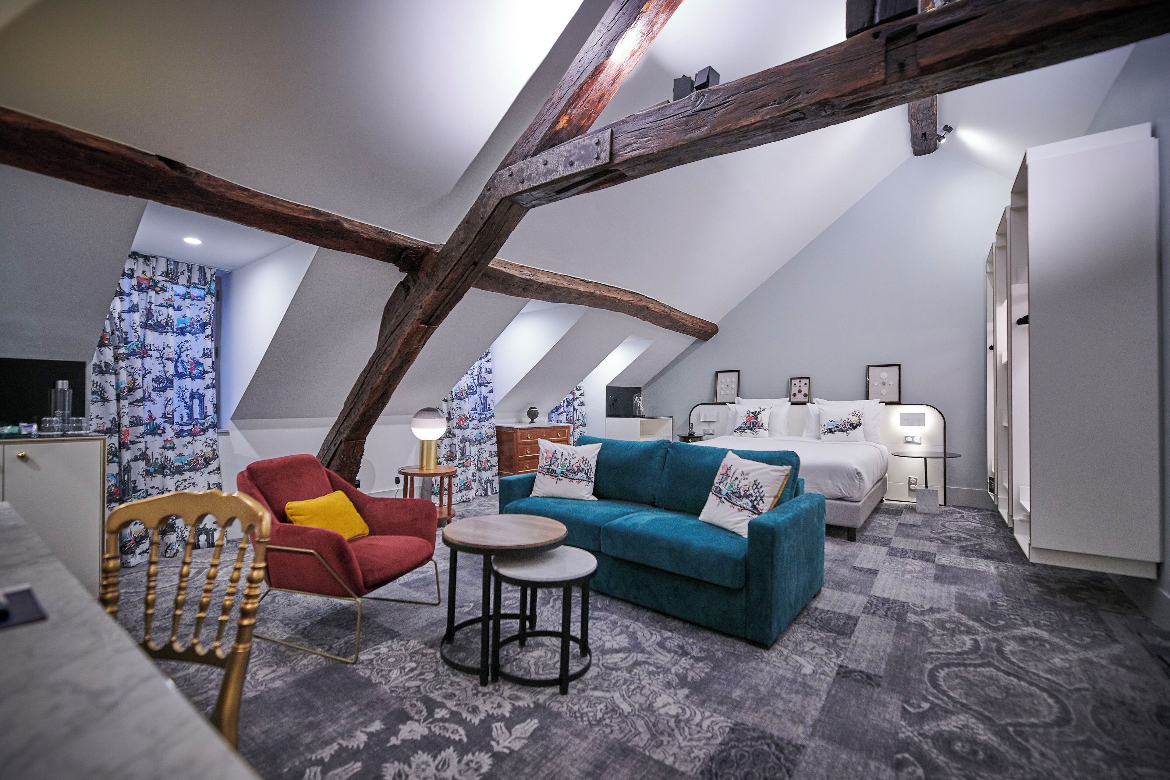 Une pièce de style « loft », avec un plafond haut, des poutres apparentes, un canapé, un fauteuil rouge, une petite table ronde et un lit avec une tête de lit blanche.