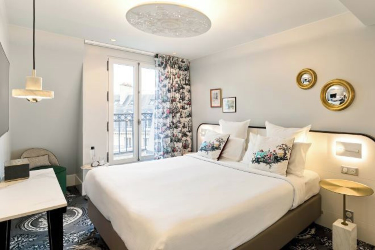 Een hotelkamer met een bed, een bureau, een stoel, een lamp, een raam en uitzicht op gebouwen.