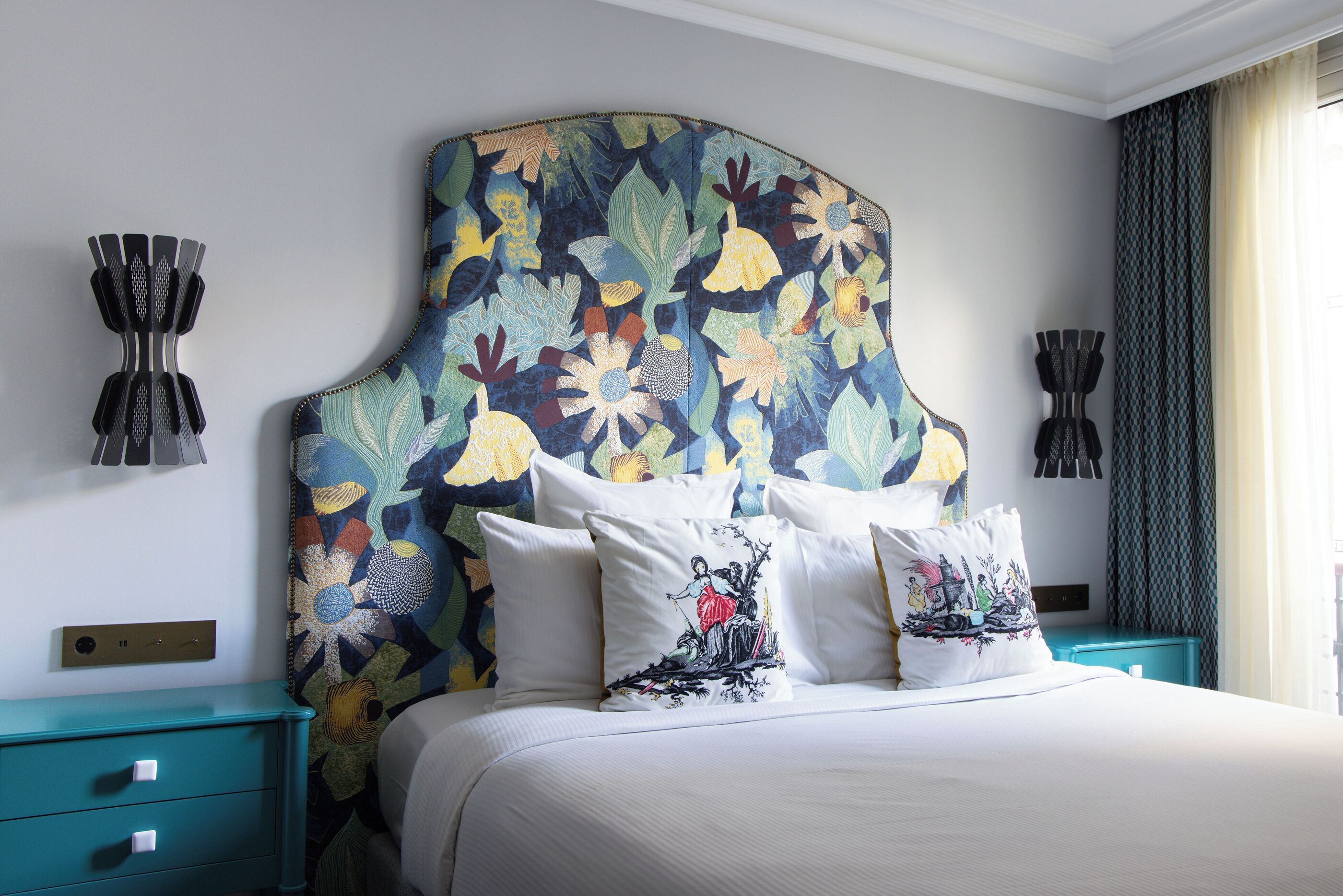Una habitación de hotel con una cama grande, ropa de cama blanca, mesitas de noche color turquesa y un cabecero estampado con motivos florales.