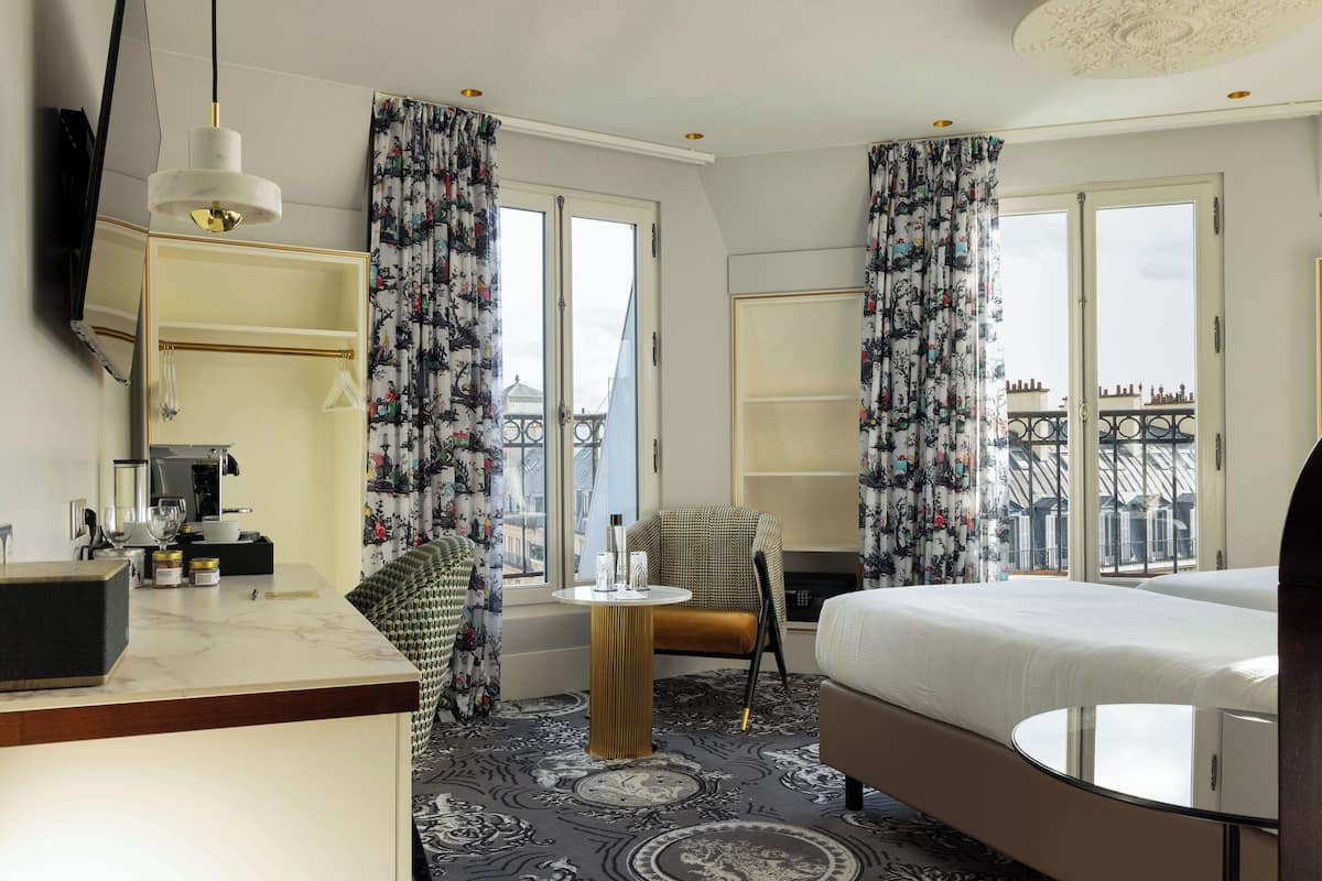 A hotel room with a bed, a chair, a small table, a mirror, and a view of the cityscape through large windows.