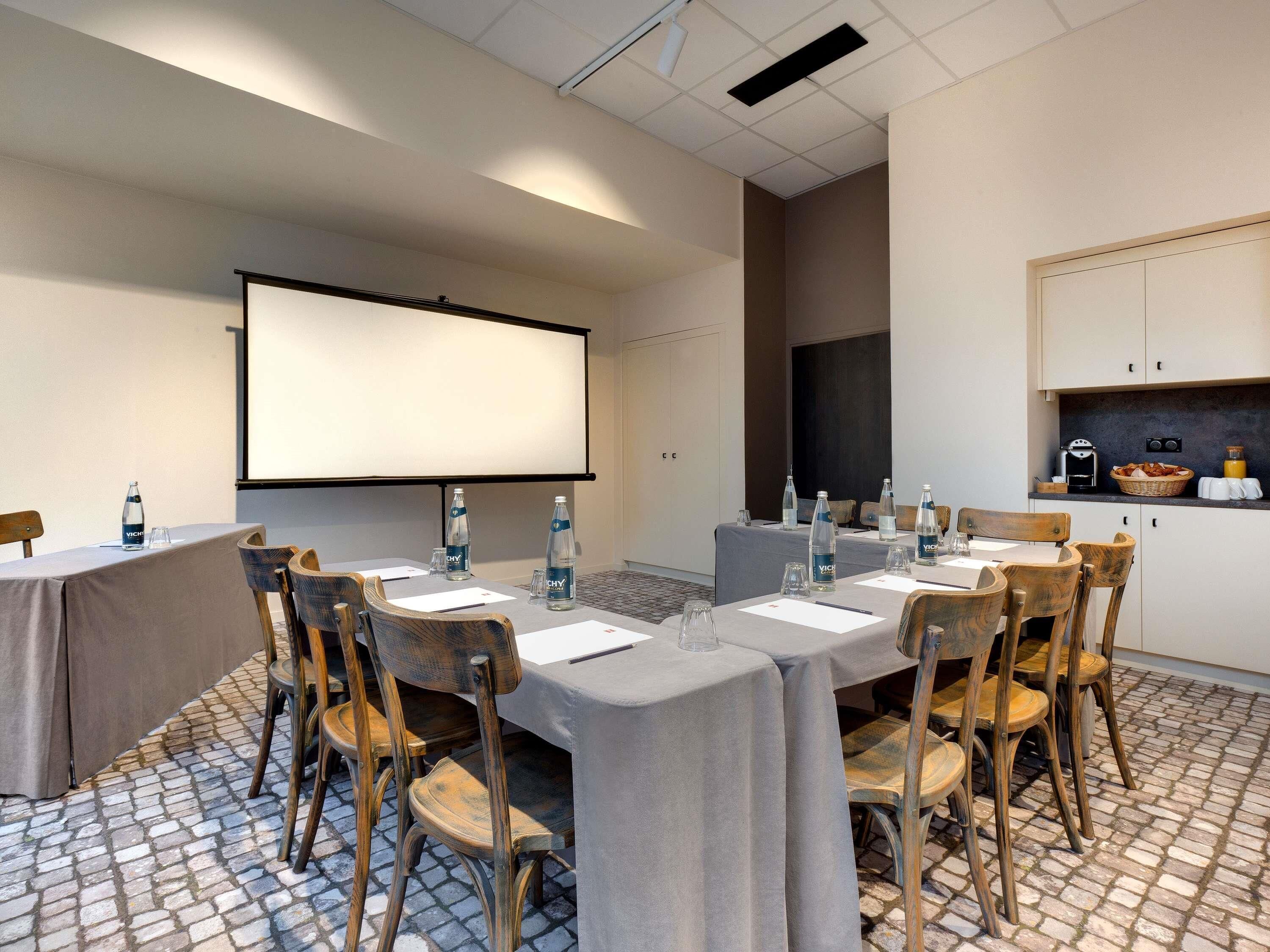 Un salón de conferencias con una pantalla grande, mesas con sillas, botellas de agua y una kitchenette.