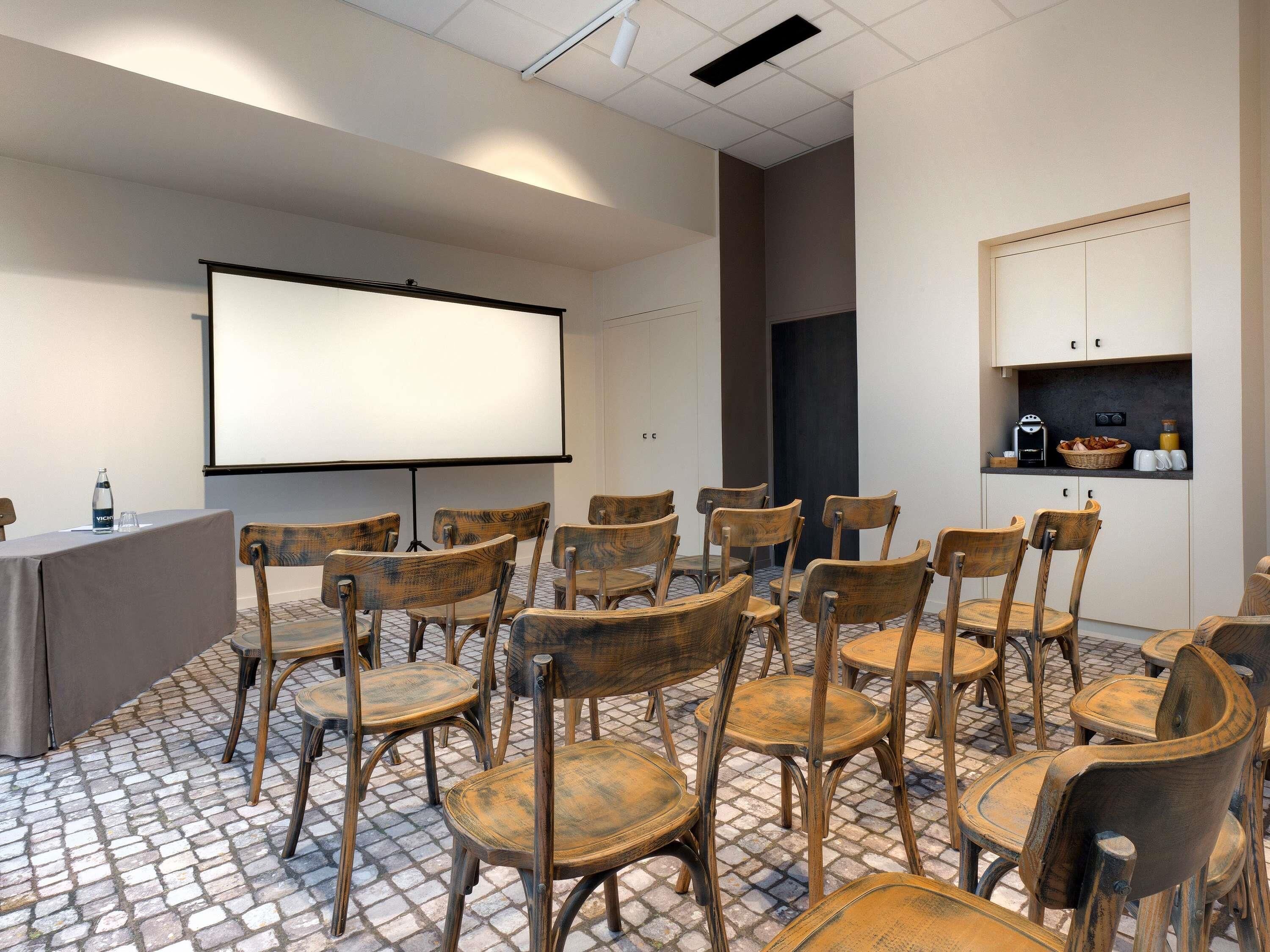 Une salle de conférence équipée d’un écran de projection, de chaises en bois, d’une table avec une bouteille d’eau et d’un coin cuisine.