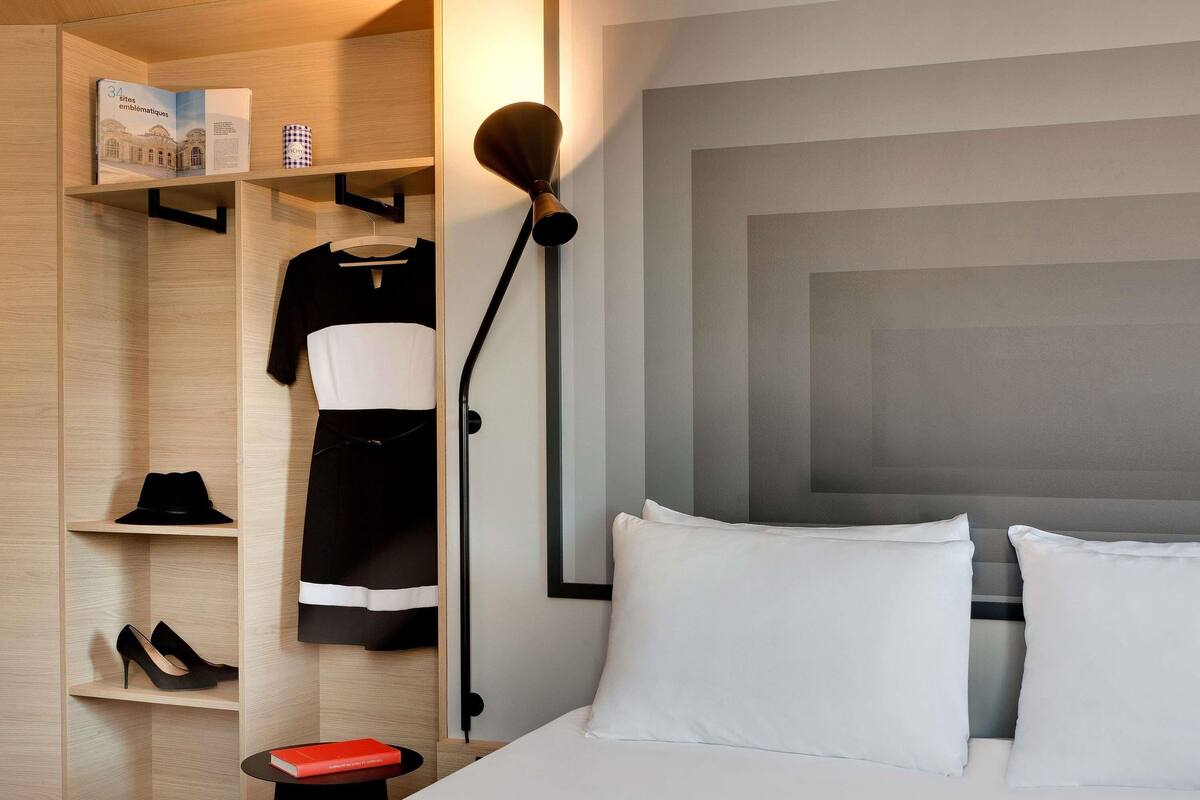 A hotel room with a bed, a wardrobe, a standing lamp, and a book on a small table.