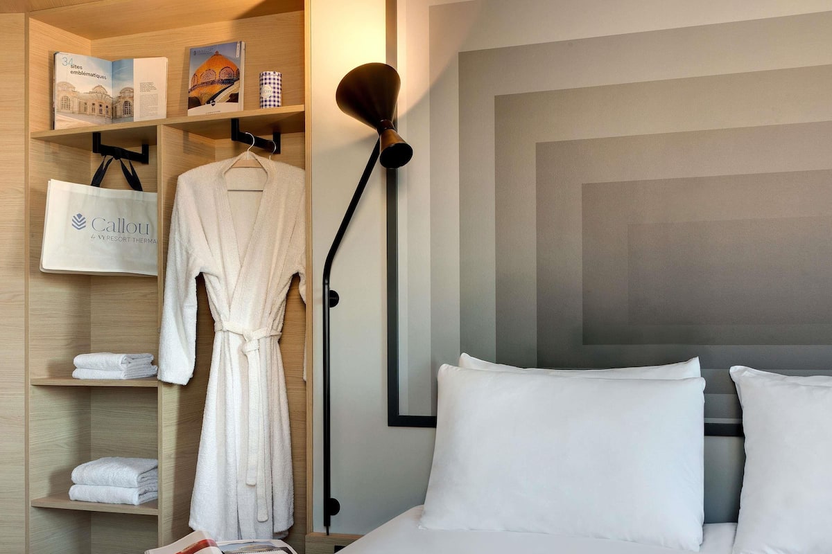 A hotel room with a bed, pillows, a robe, a towel, a magazine, a lamp, and a shelf with a book and a bag.