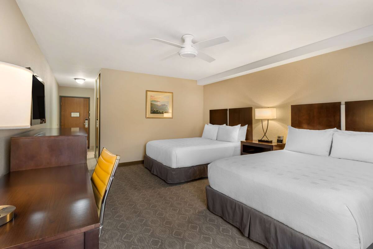 A hotel room with two beds, a desk, a chair, a ceiling fan, and a painting on the wall.