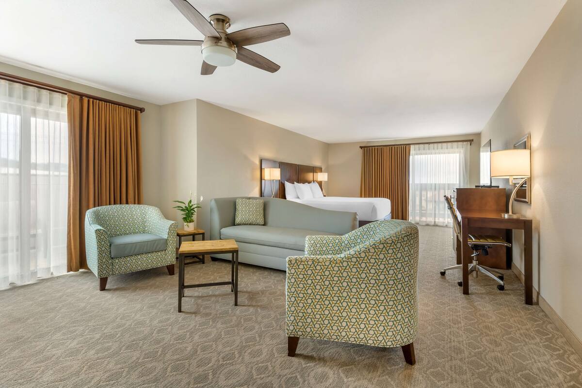 A hotel room with a bed, two armchairs, a desk, a ceiling fan, and a large window with curtains.