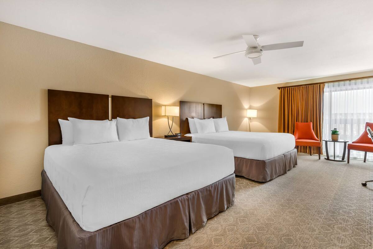 A hotel room with two beds, a ceiling fan, a small table with a plant, and an orange chair.