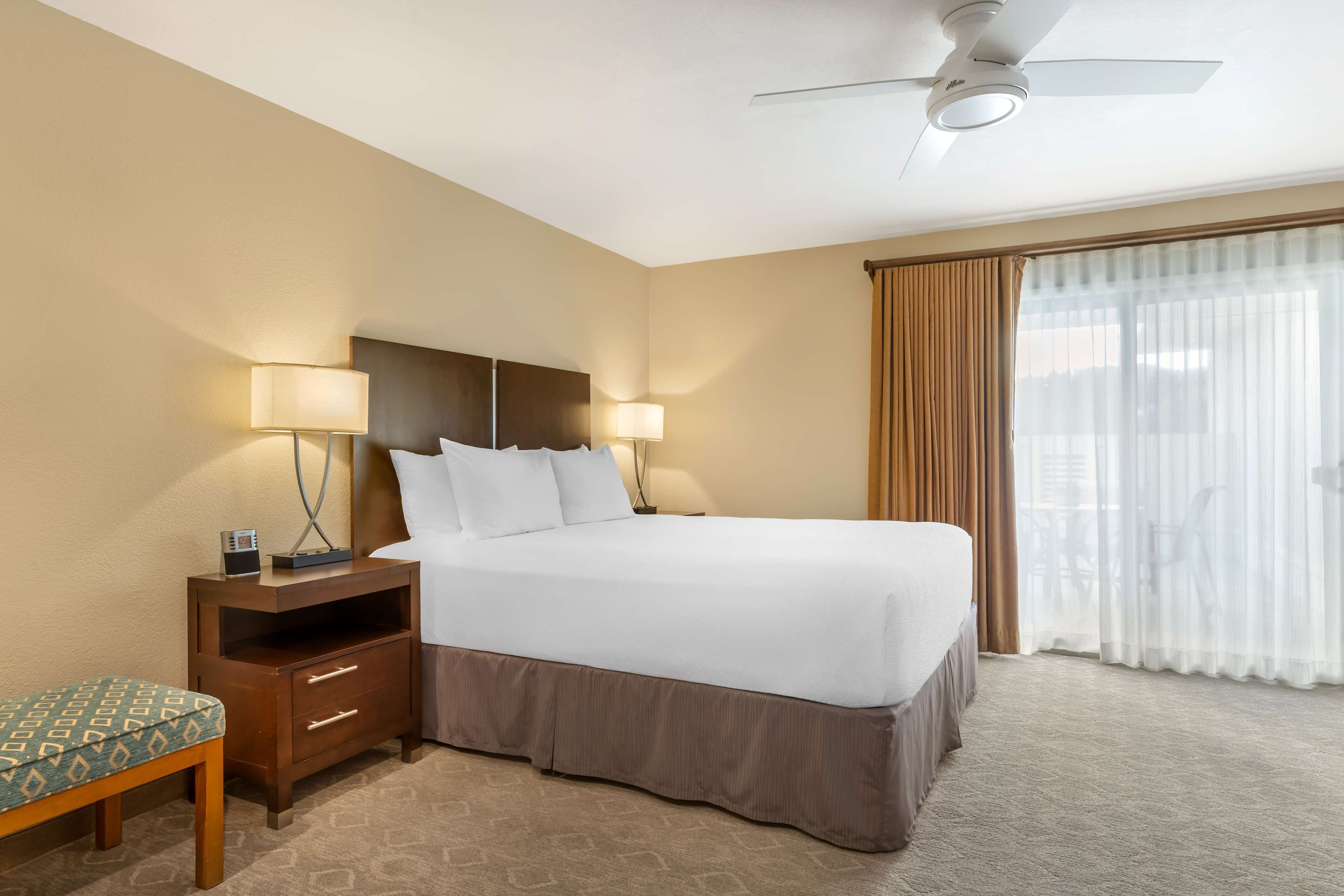 A hotel room with a bed, bedside lamps, a nightstand, a chair, and a view of the outdoors through a window.