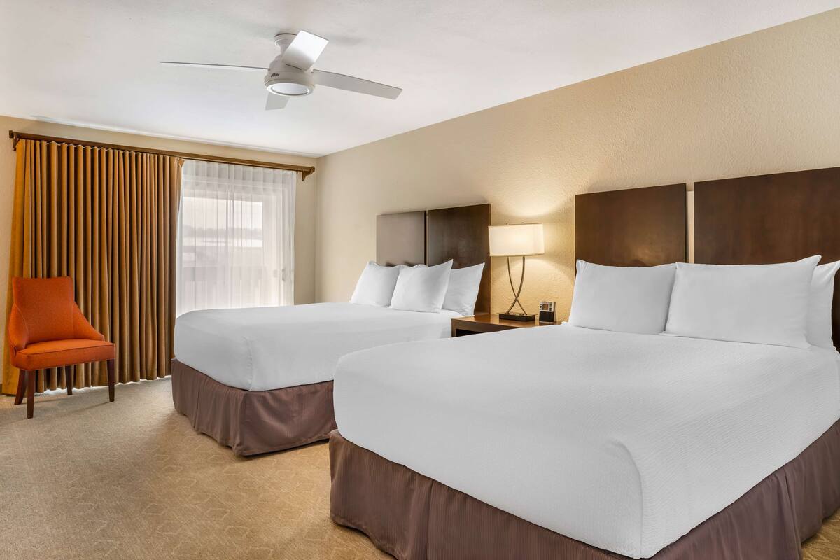 A hotel room with two beds, a ceiling fan, a chair, and a window with curtains.