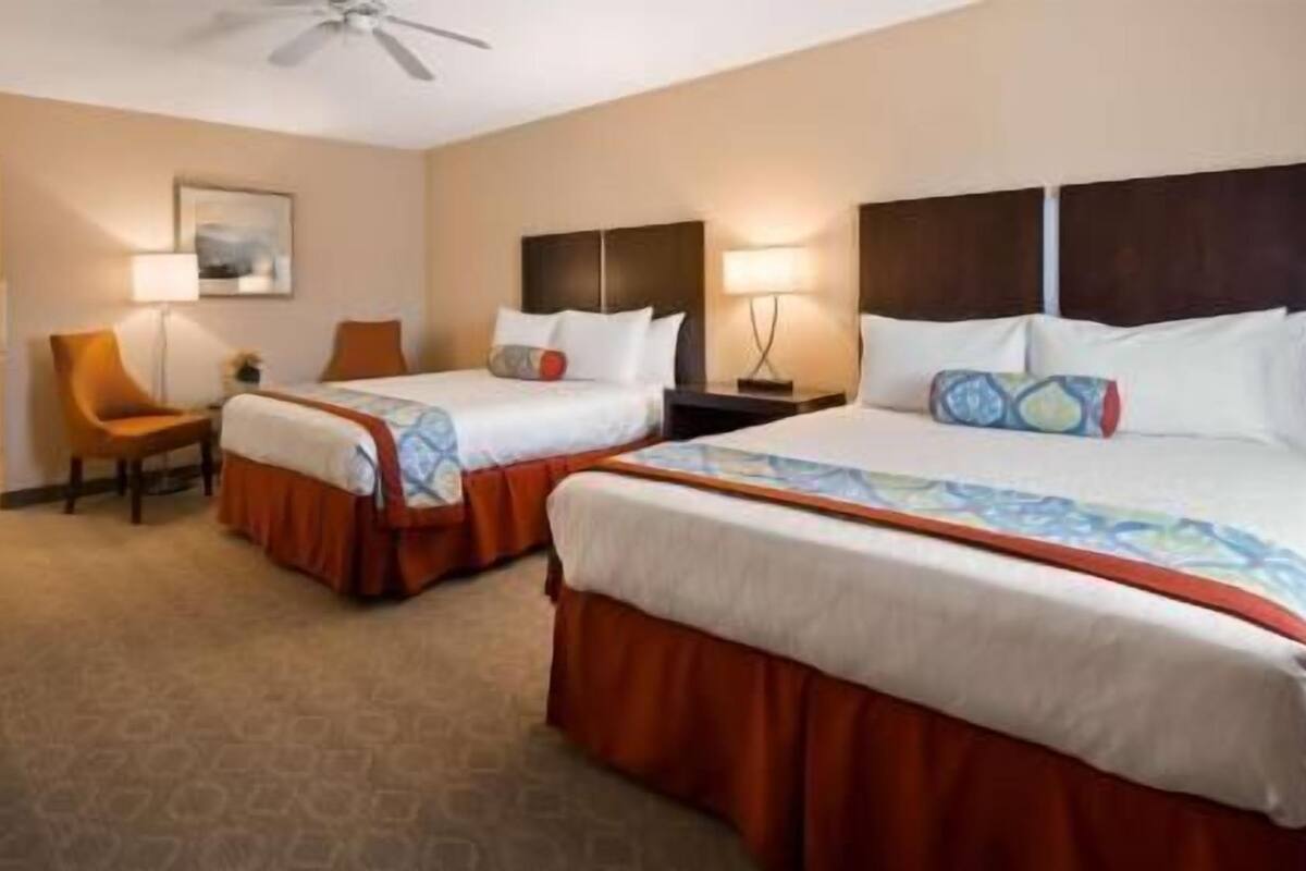 A hotel room with two beds, a chair, a desk, and a ceiling fan.